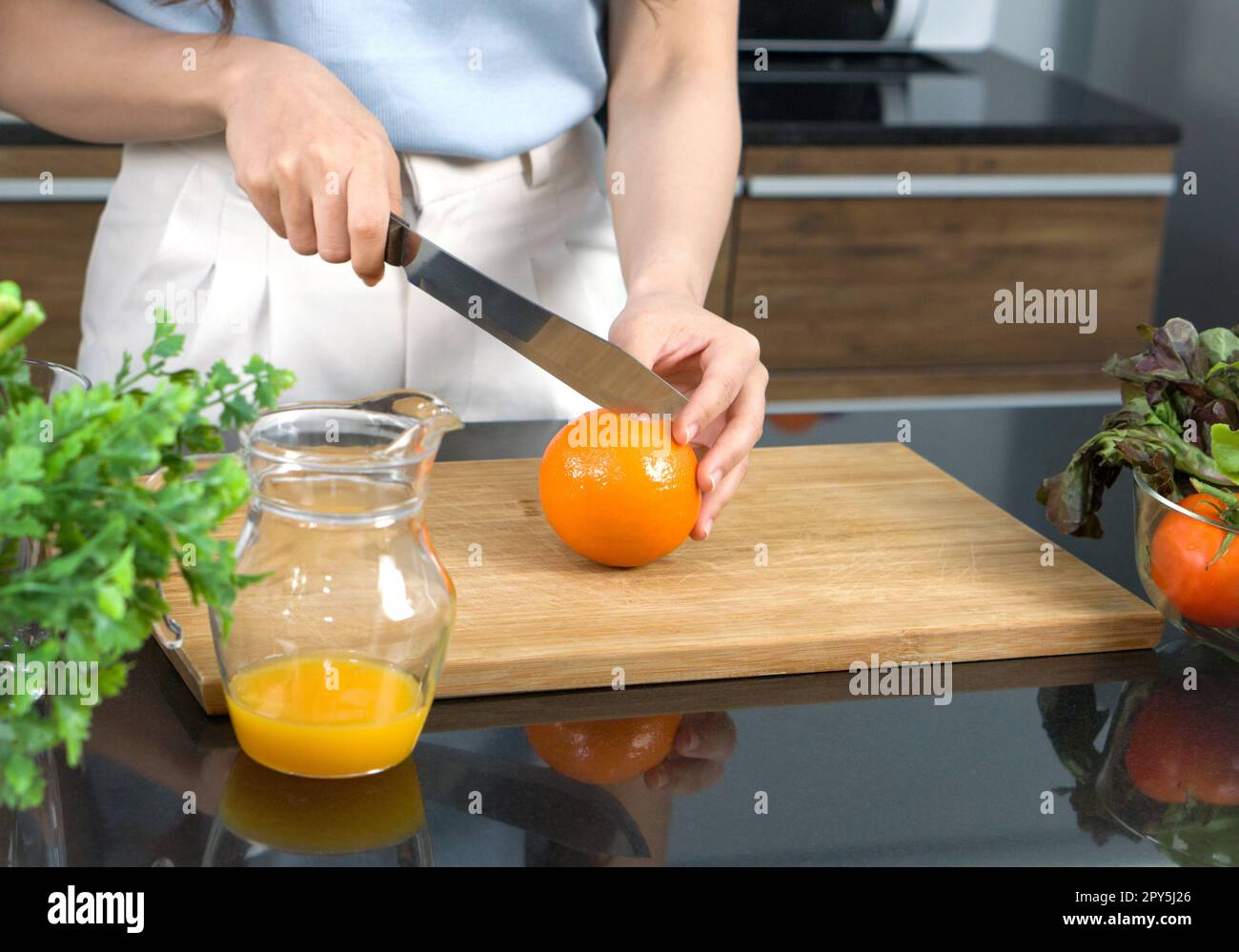 Closeup hand holding knife cutting orange fruit on a wooden chop board. Jar with mixed fruit juice  placed on the kitchen counter. Stock Photo
