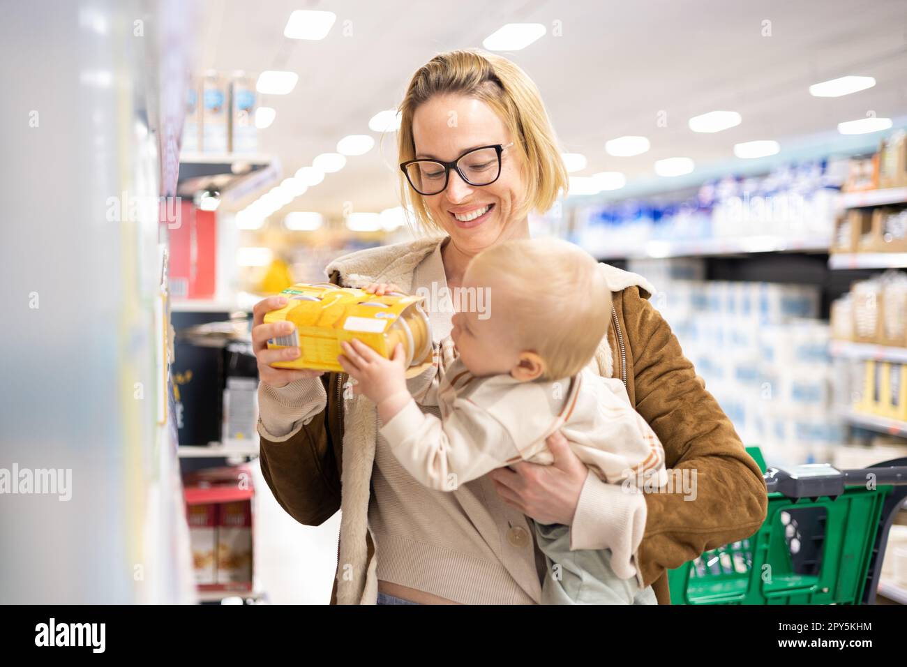 Caucasian mother shopping with her infant baby boy child choosing products in department of supermarket grocery store. Stock Photo