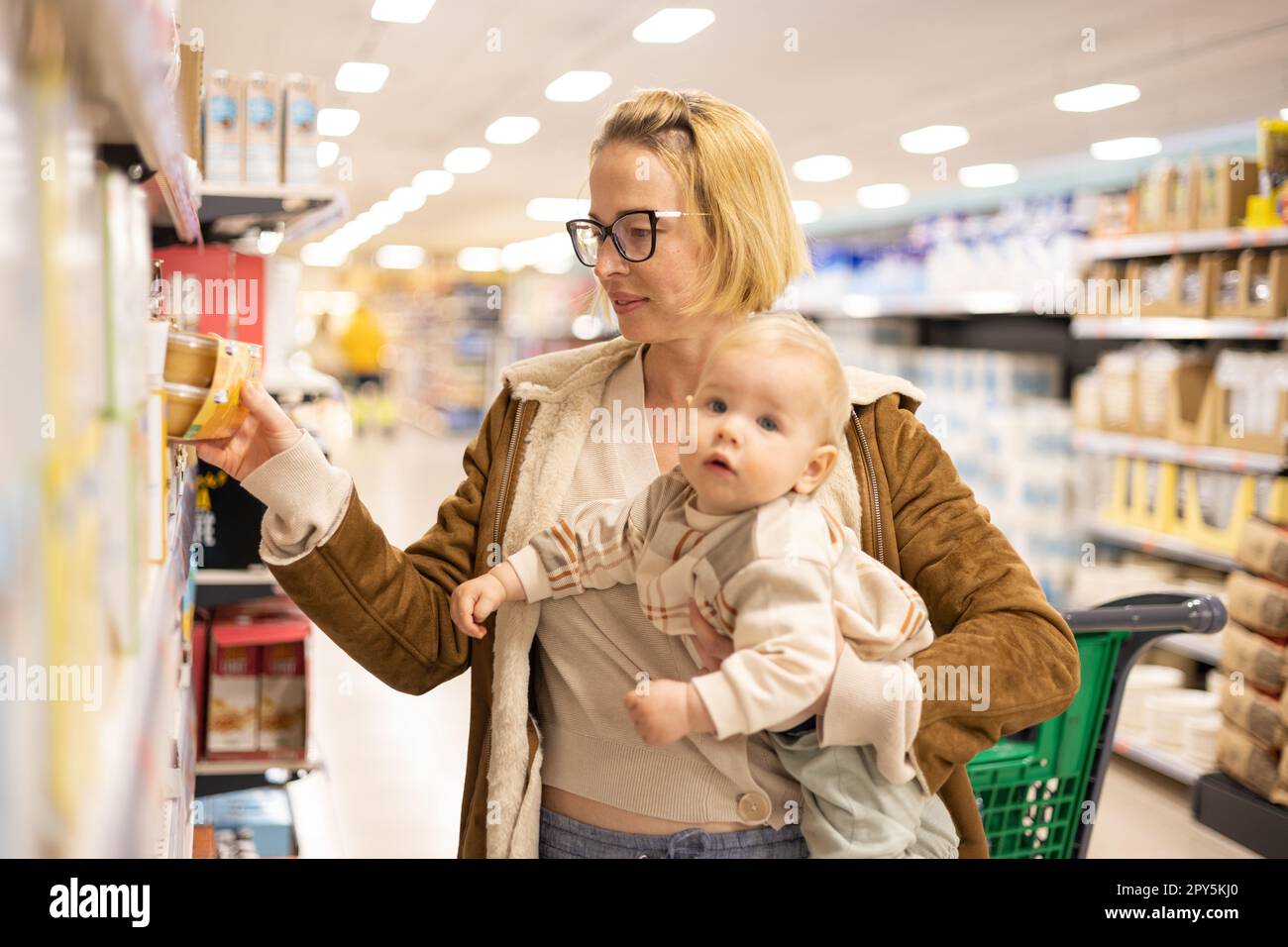 Caucasian mother shopping with her infant baby boy child choosing products in department of supermarket grocery store. Stock Photo