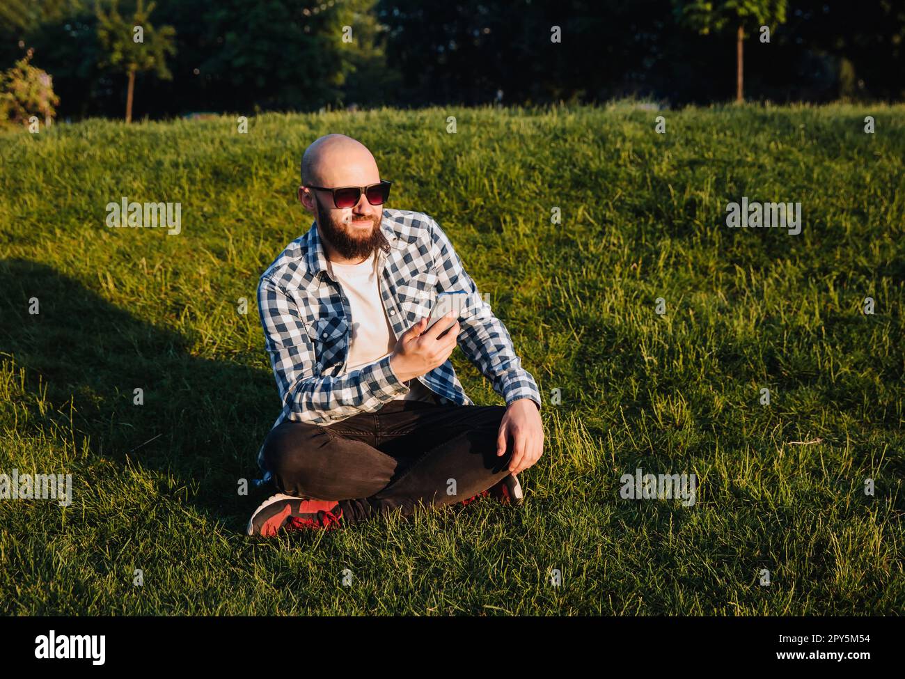 A young man sits on the green grass in the park and uses social networks using a mobile phone. Stock Photo