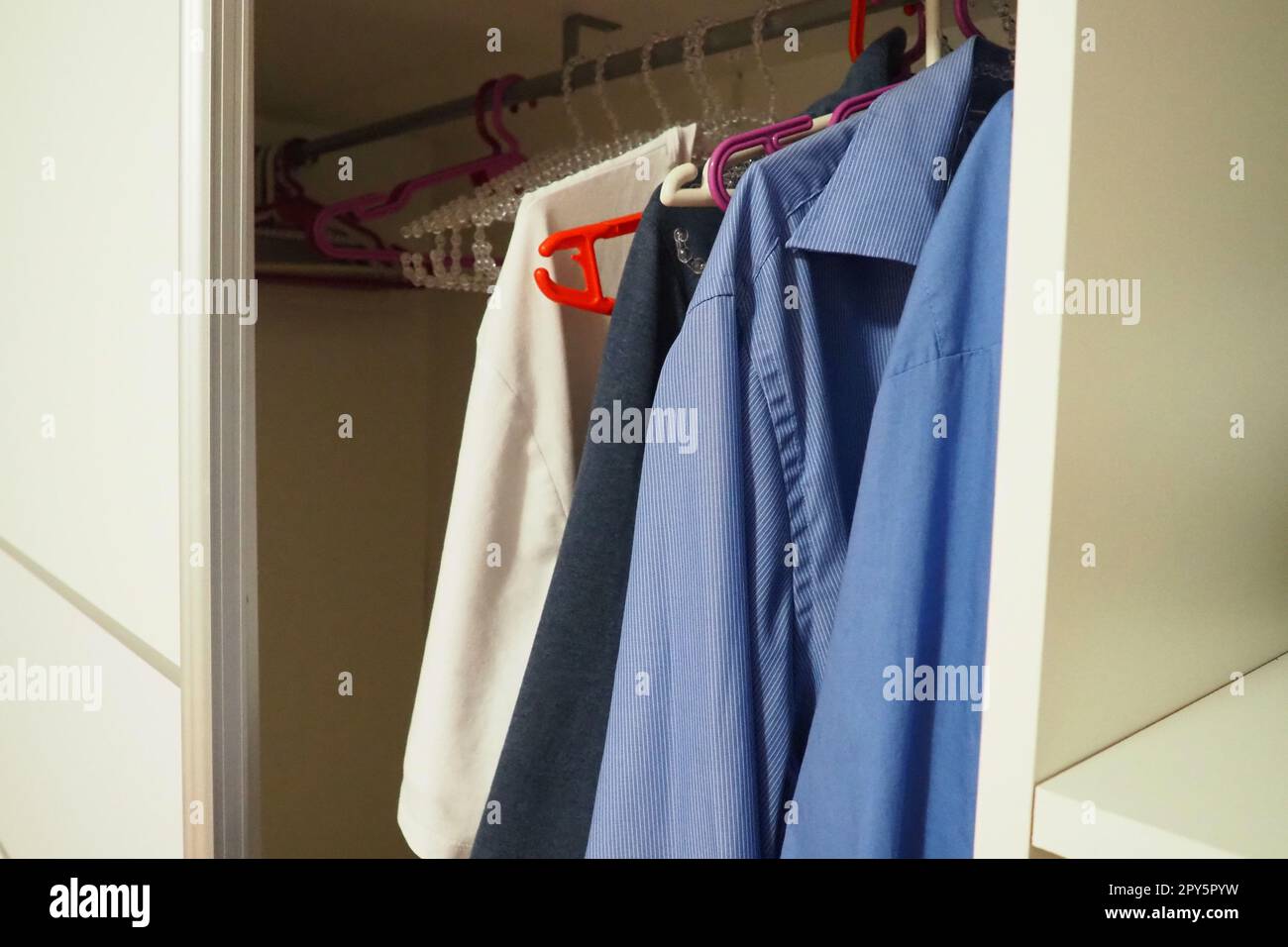 Men's shirts hang on hangers in an open white closet. Men's fashion. Organization of things in a closet or dressing room. Blue and white shirts. Housekeeping. Stock Photo