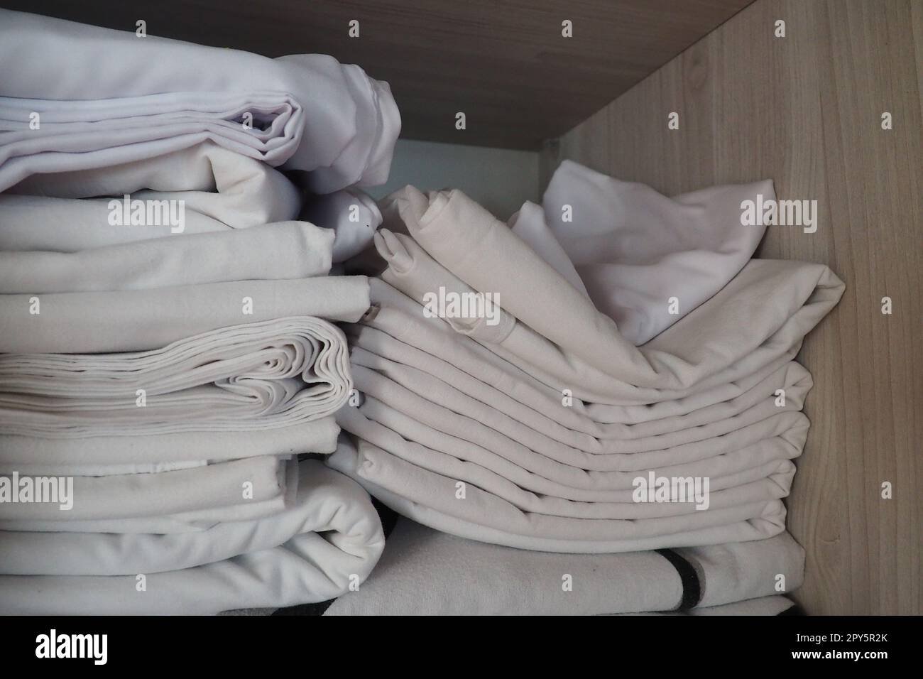 The white linens are stacked in the closet. Internal contents of a linen closet. Household organization. Hygienic conditions for sleep. Fabrics and bed sheets in half open white closet or wardrobe Stock Photo