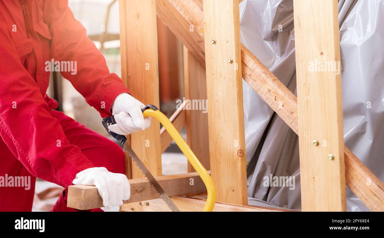 A worker is sawing wood to assemble a crate for moving an industrial machine. A woman in red mechanic coveralls hand holding a bow saw cutting the wooden plank of a crate. Safety in workplace concept. Stock Photo