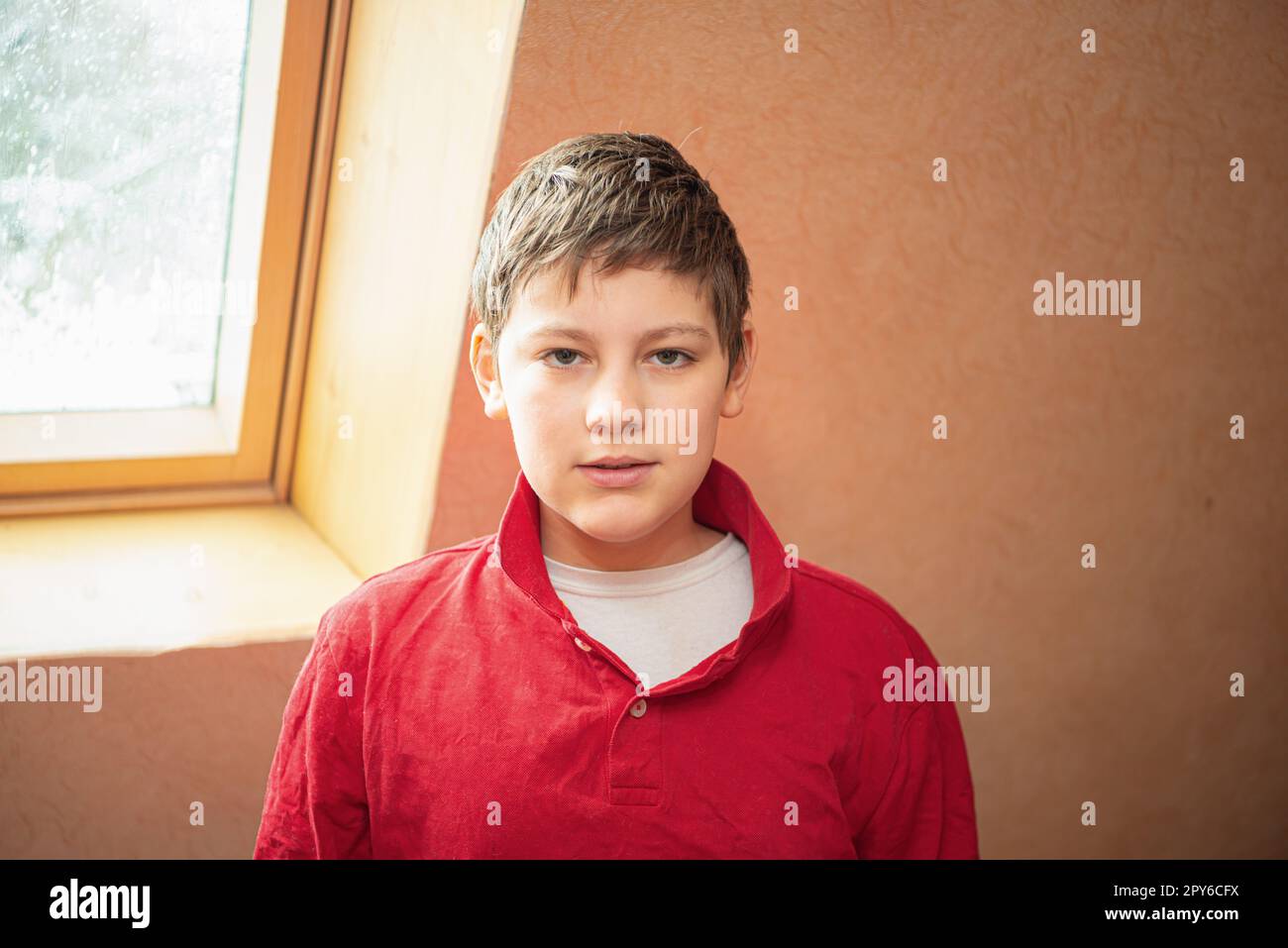 A white boy of school age stands near the window, dressed in a red shirt. Stock Photo
