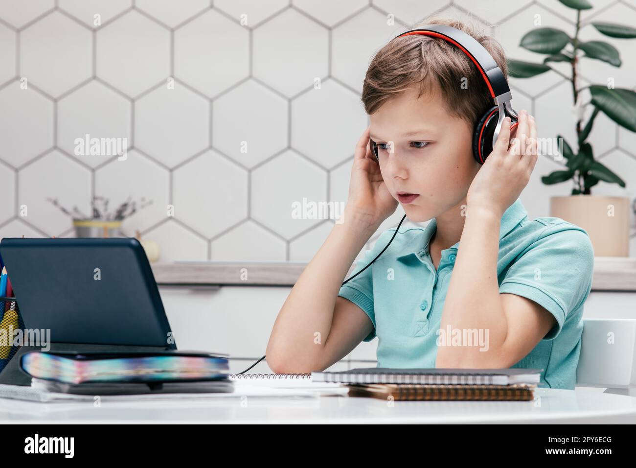 Portrait of young preteen boy covering headphones with hands, looking at screen of tablet, listening to online lesson. Stock Photo