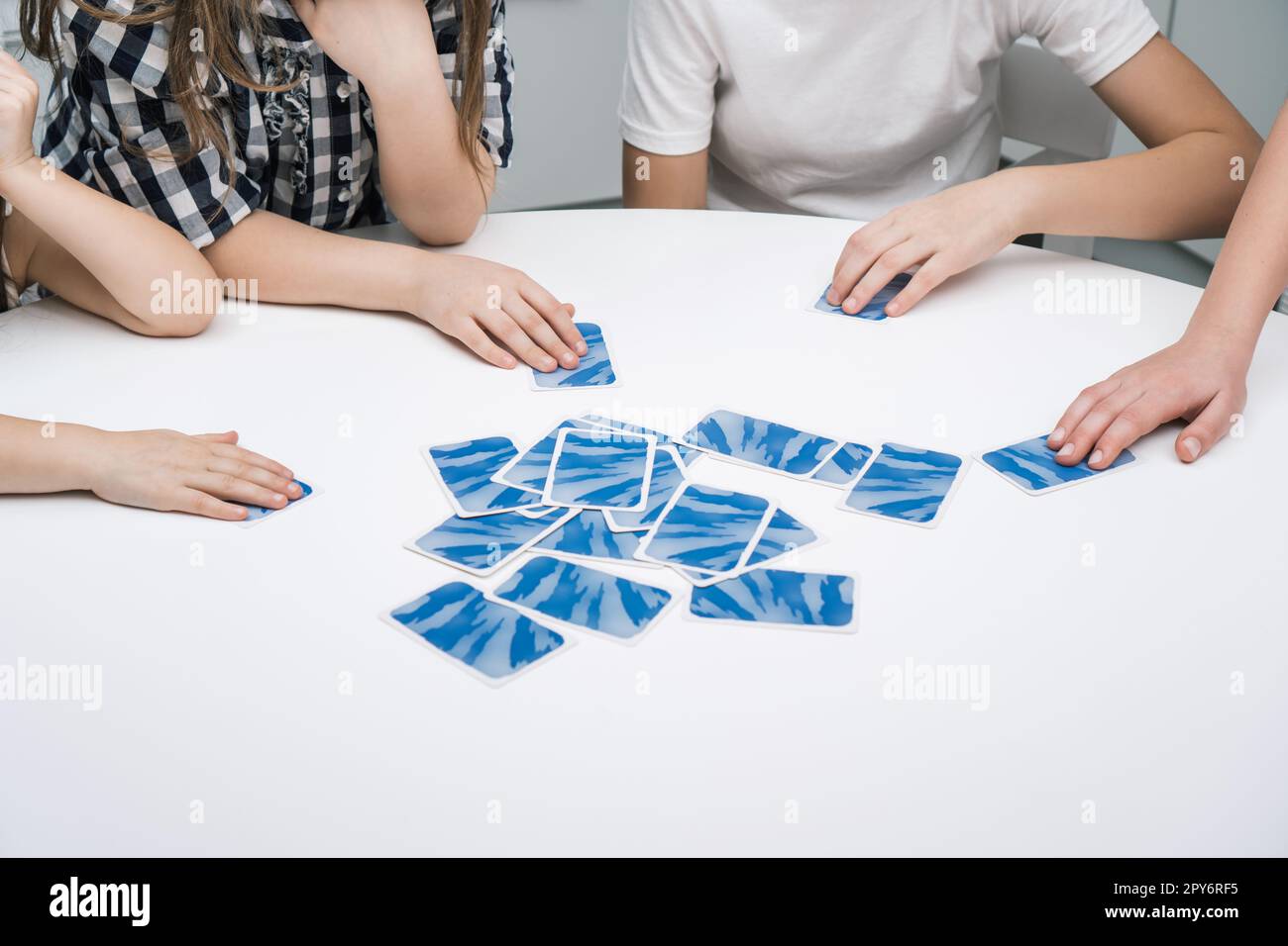 Group of friends play card game sitting at white table. Children take with hands flipped paper blue cards from pile. Stock Photo