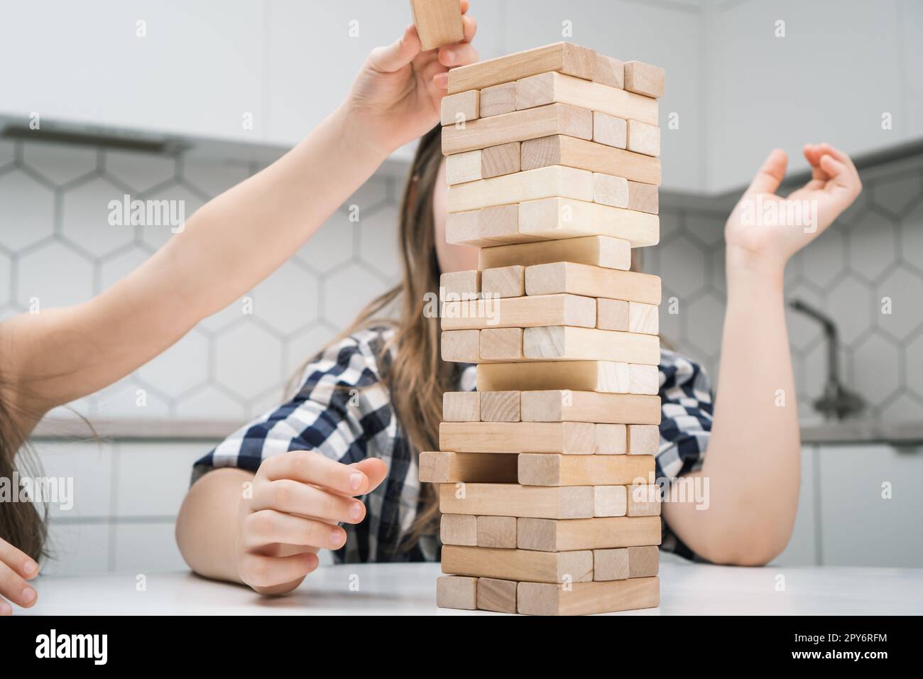 School kids play jenga sitting at kitchen table closeup. Hand of girl put wooden block on top of tower keeping balance. Stock Photo