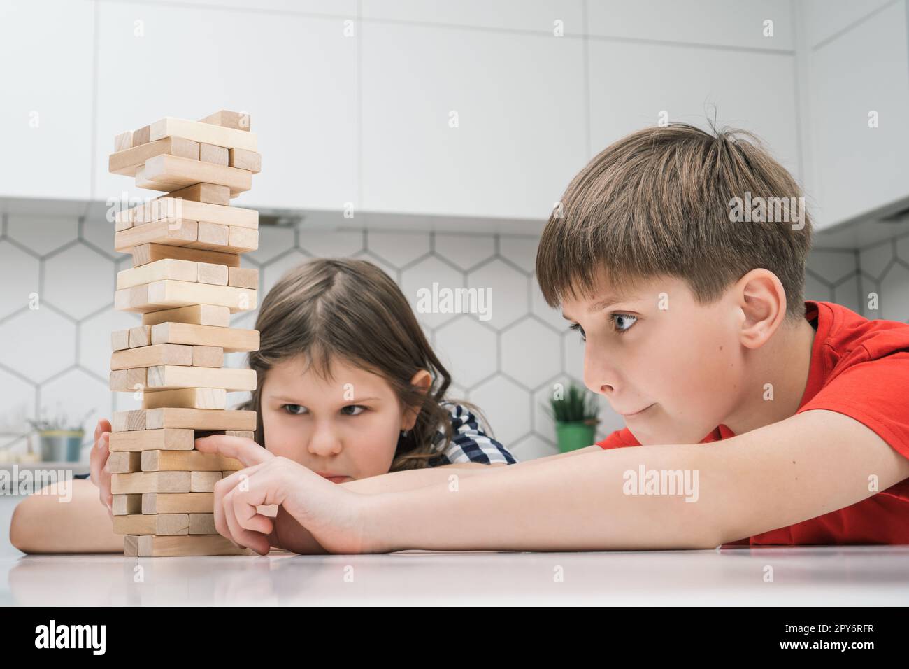 Happy school kids play tower sitting at kitchen table. Concentrated boy and girl build tower from little wooden blocks. Stock Photo