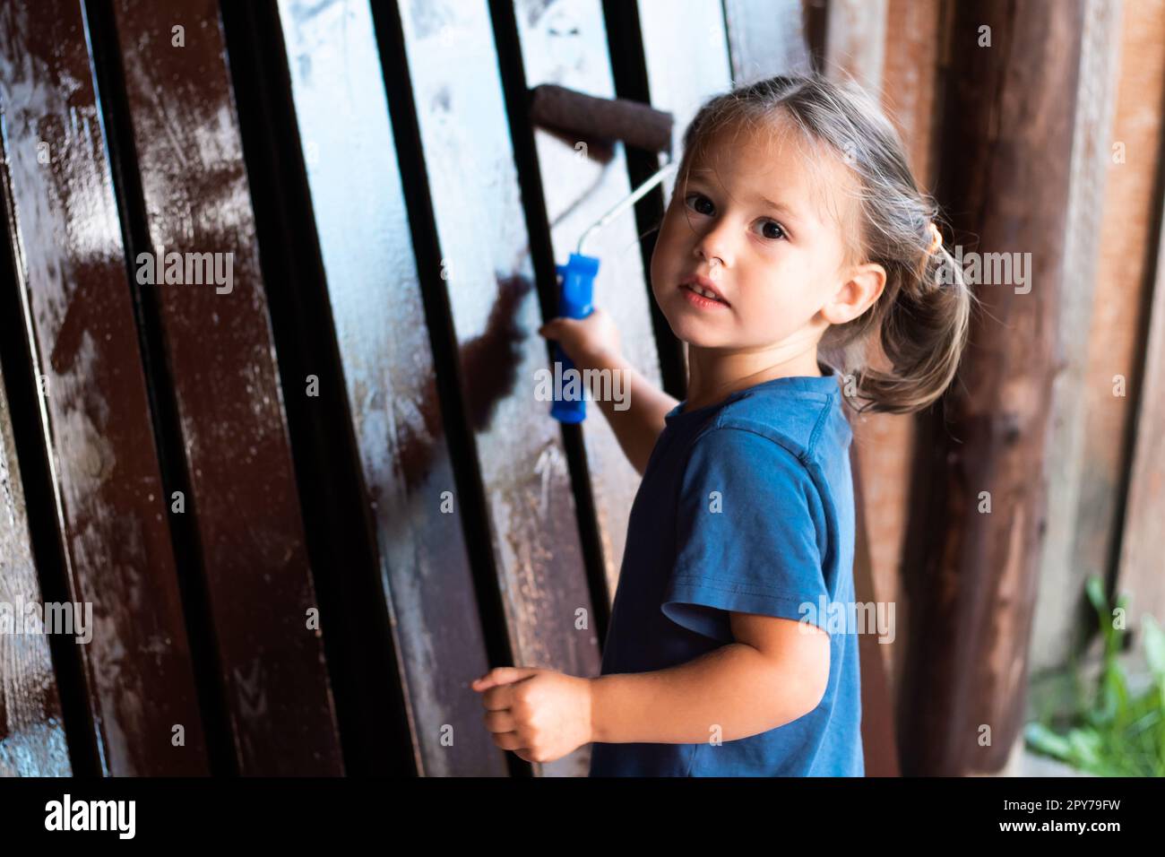 Cute blonde little girl carry out painting work, coloring wooden fence border with paint roller and looking at camera Stock Photo
