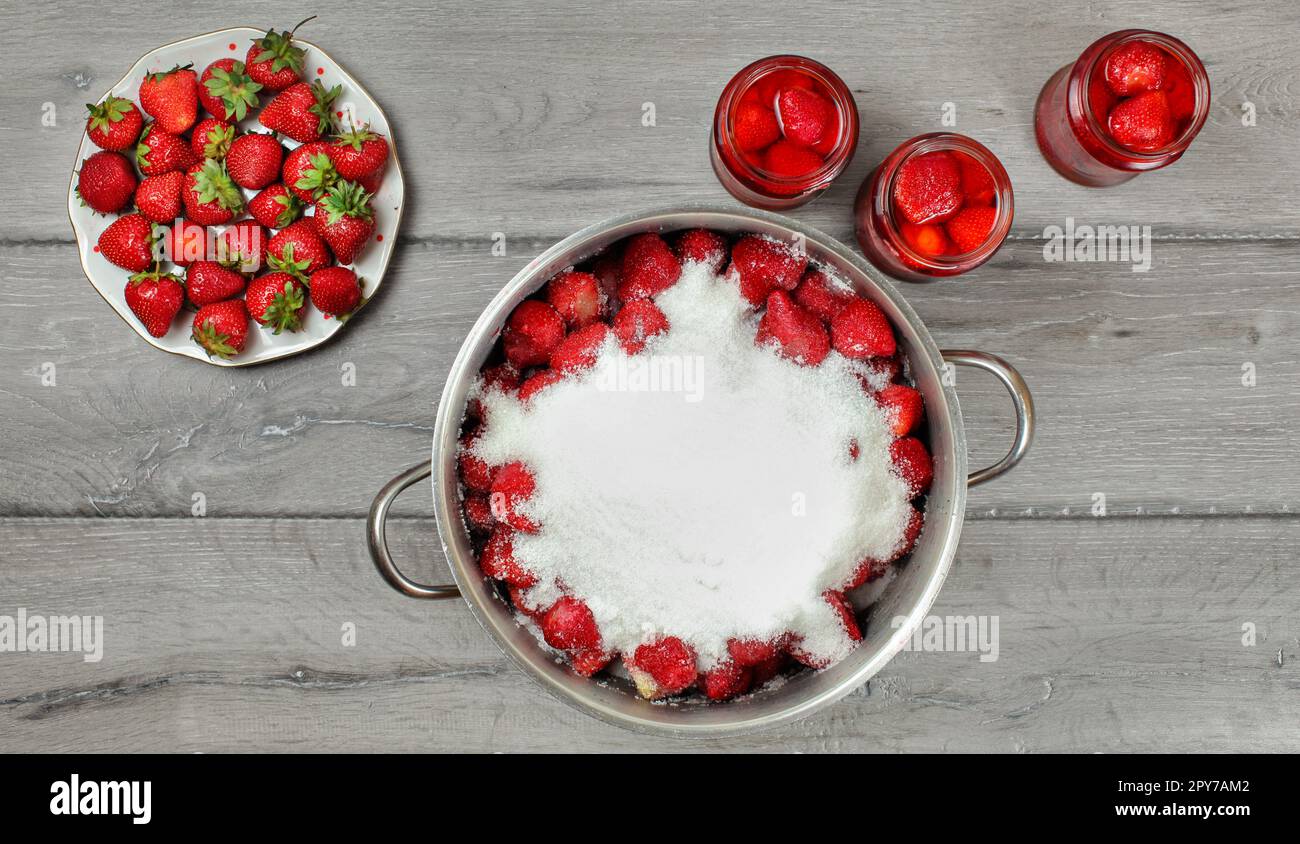 Tabletop photo - large steel pot of strawberries covered with crystal sugar, plate and glass bottles with more fruits around on gray wood desk. Stock Photo