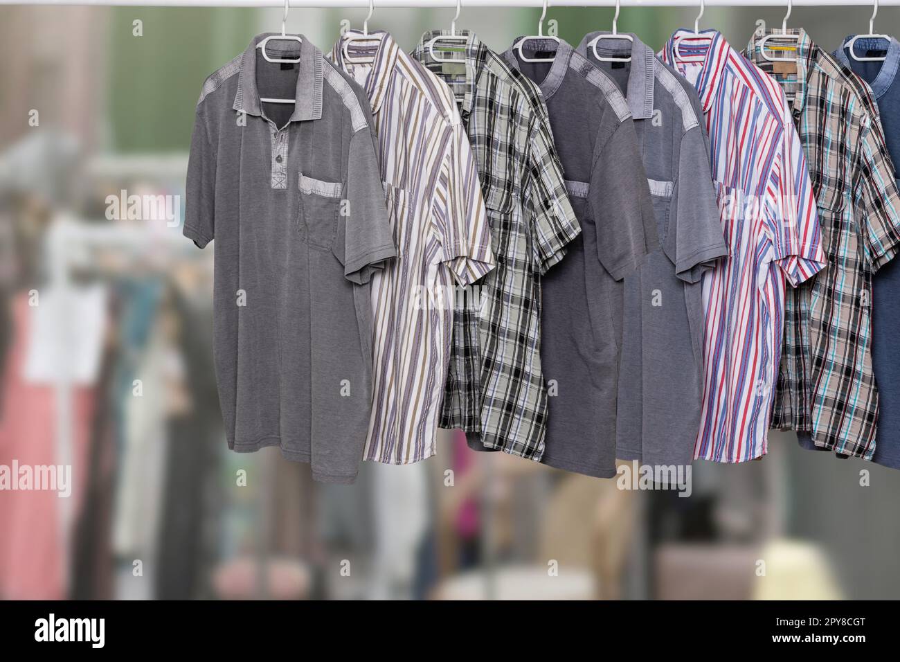 Clothes on rack. Clothing rack with various colorful male casual summer shirts over abstract blurred clothes shop. All hanging neatly on hangers at cloth rail. Space. Shopping. Stock Photo
