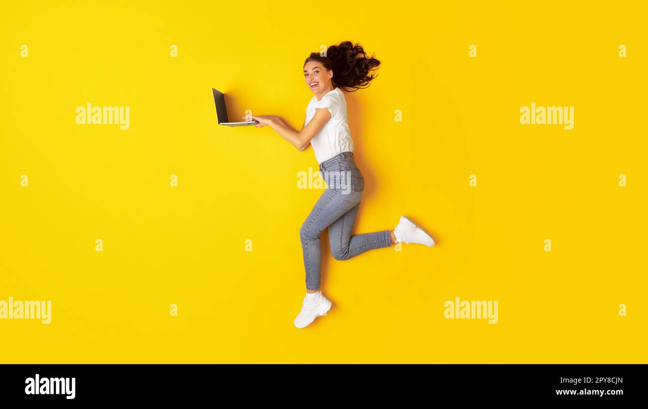 Cheerful Young Woman Using Laptop Computer Jumping Over Yellow Background Stock Photo