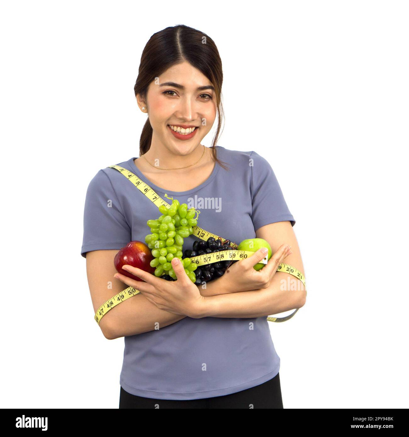 Young asian woman in fitness clothes holding fruits with  measuring tape. Portrait on white background with studio light. Healthy nutrition and weight losing concept. Stock Photo