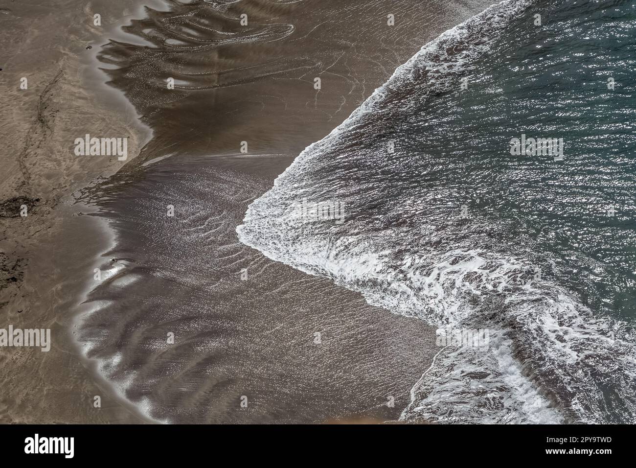 View of the beach with dark sand and the sea water, water with white foam and the effect of the sun reflecting in the water, Madeira Island, Portugal. Stock Photo