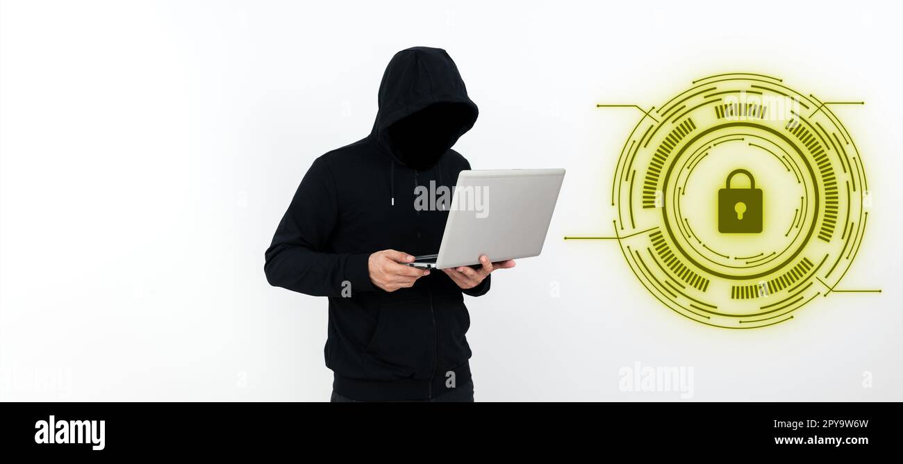 Male programmer in black hoodie stands and holds laptop. Hacker with hood on his head typing text on computer. Futuristic digital design on the wall. Stock Photo