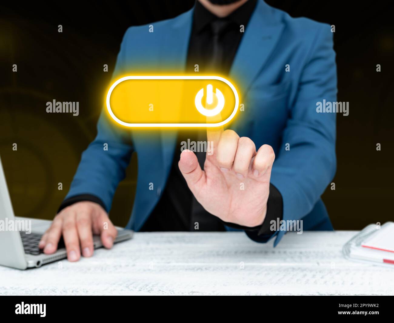 Businessman in blue jacket sitting at the white table and pressing virtual button. Laptop laying on the desk.Futuristic style image with colored glow. Stock Photo