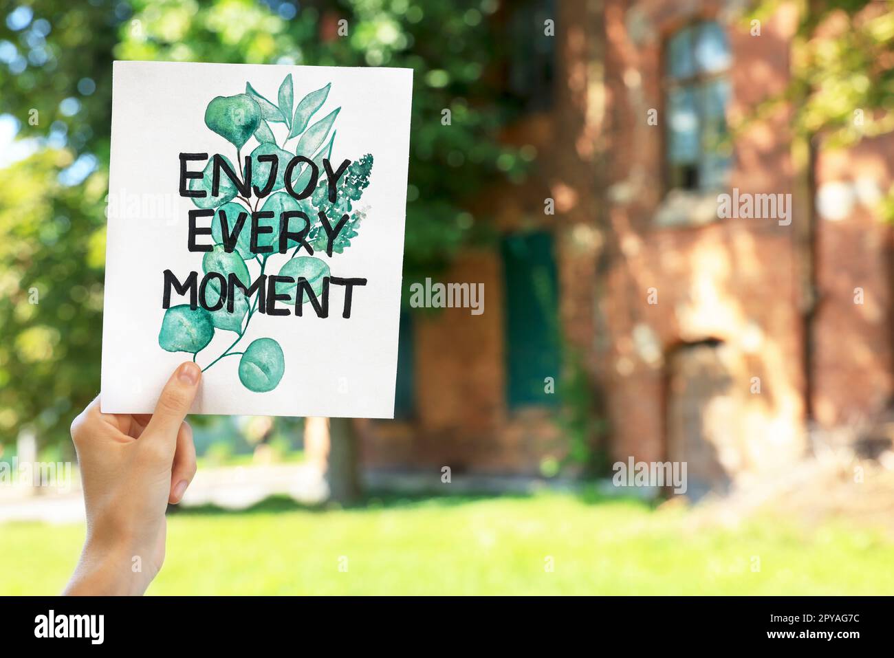 Woman holding card with phrase Enjoy Every Moment outdoors, closeup. Space for text Stock Photo