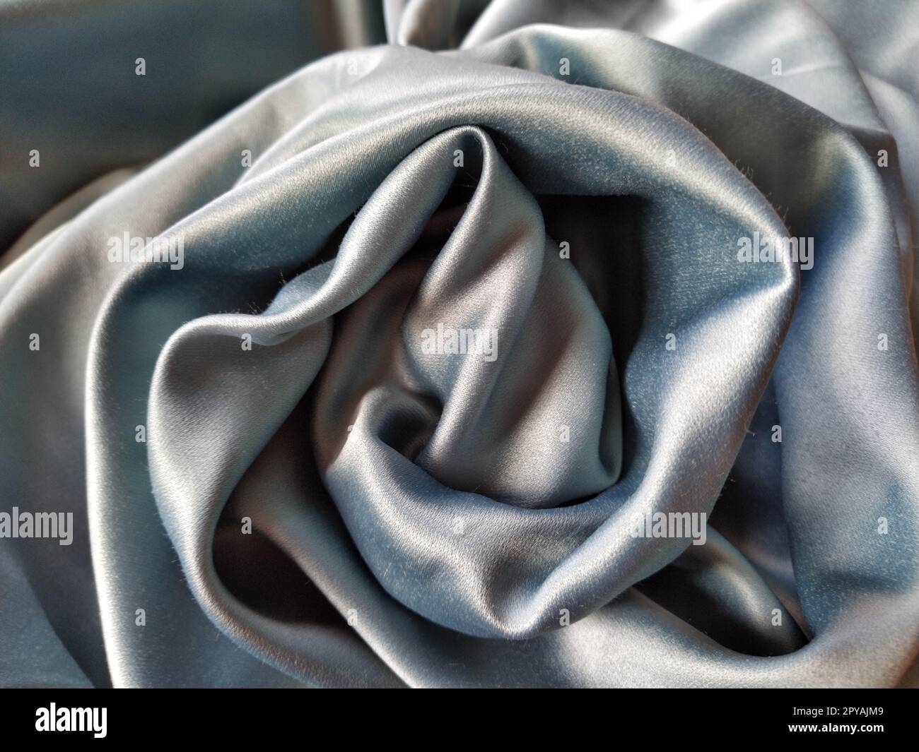 Silky to the touch fabric of gray - blue color with a brilliant shimmer. Natural lighting, light and shadow. The material is casually folded into a circle, resembling a rose bud. Polyester Textile. Stock Photo