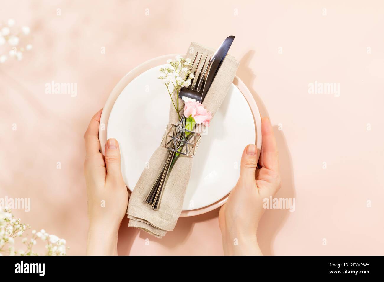 Female hands hold holiday dishes on light pink background with spring white flowers and shadows. Romantic holiday table settings. Flat lay Stock Photo