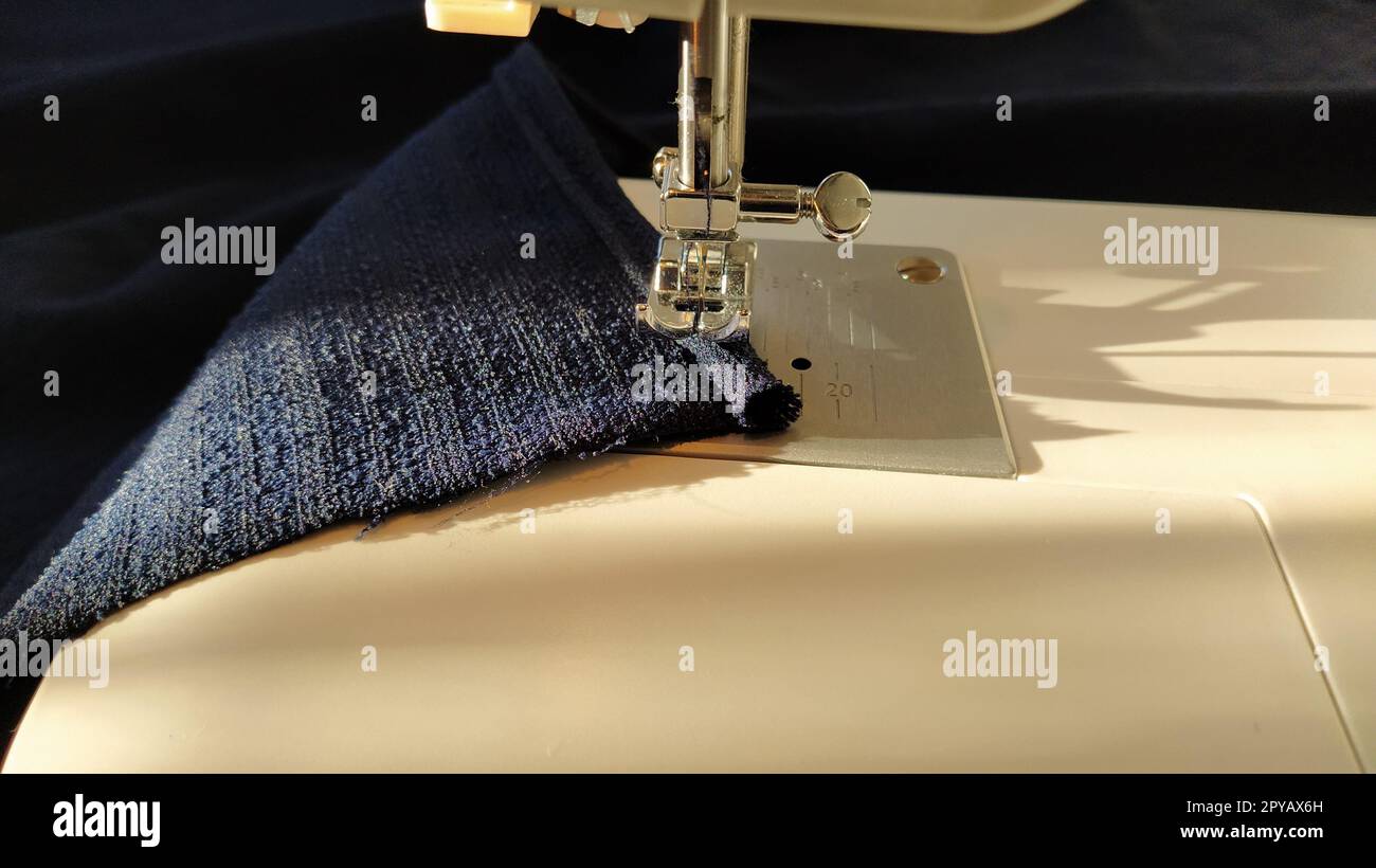 Sewing process on a modern sewing machine, close-up. Sewing machine foot with a needle. Dark blue fabric is trimmed with double wrapping of the fabric edge. Natural sunlight Stock Photo