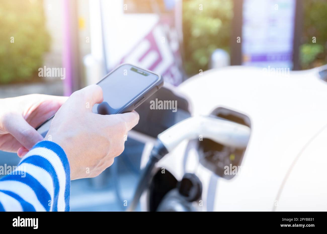 Woman using smartphone paying service in mobile app on blur EV car charging at electric vehicle charging station. EV car charging point. Commercial EV car charging station. Sustainability lifestyle. Stock Photo