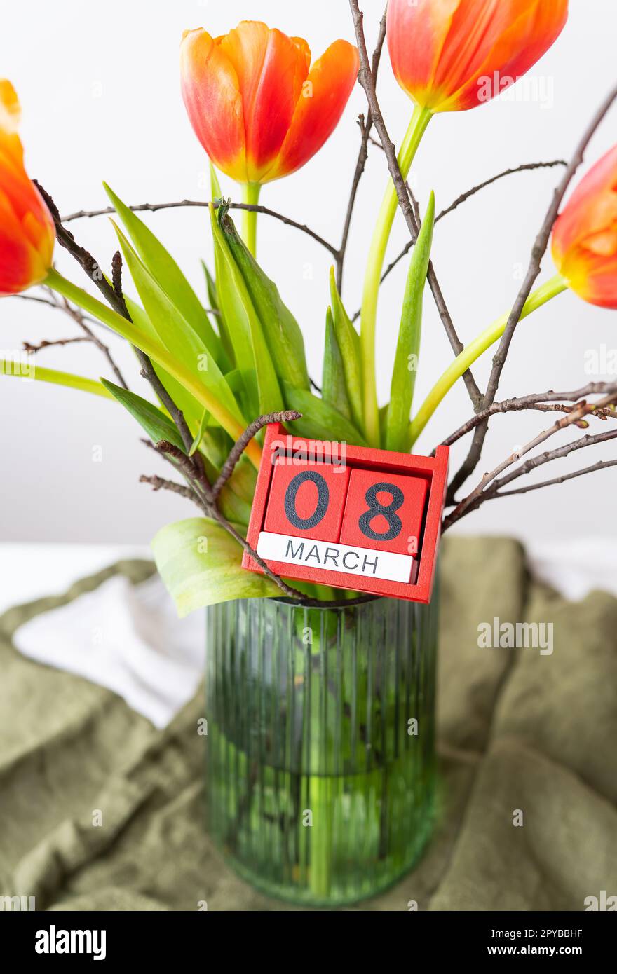 A very beautiful spring bouquet in a green vase stands on a table on a linen tablecloth, orange peony tulips. Wooden cubes with the inscription March 8. Stock Photo