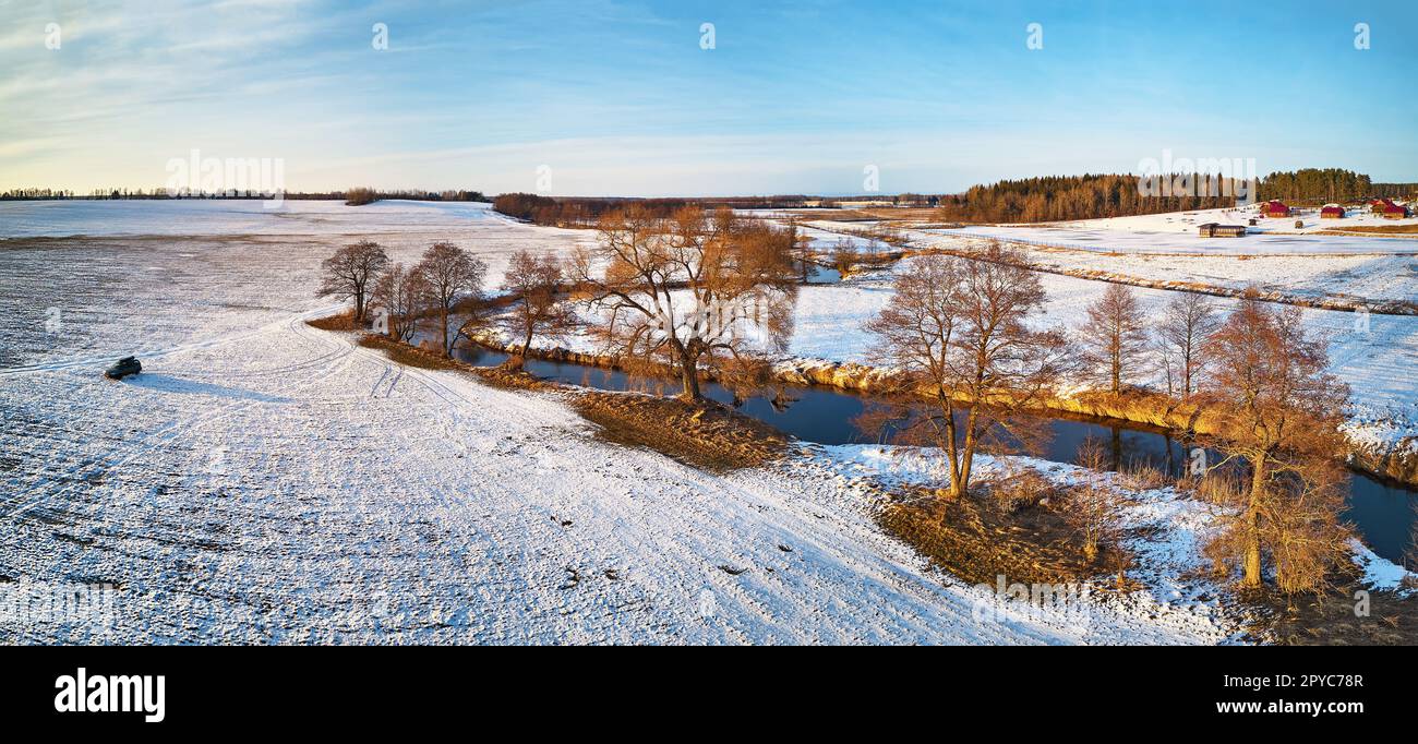 River flowing in snowy winter fields in wilderness at sunset. Stream meandering frosty countryside on cold evening. Aerial view of trees on riverbank. Stock Photo