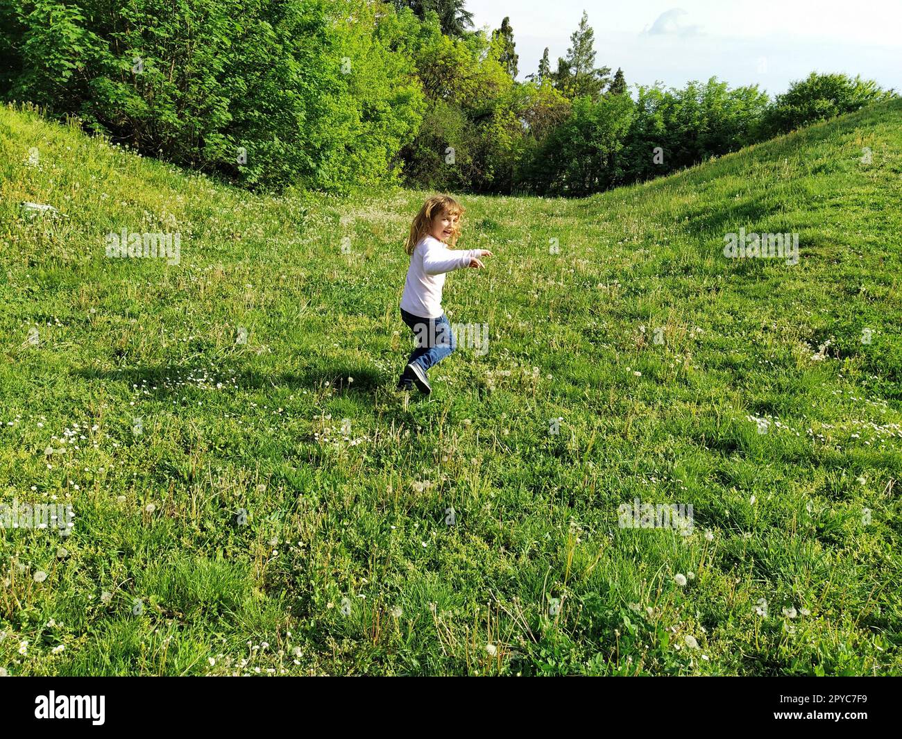 Girl runs across the field with trimmed green grass. the child is dressed in a white blouse and blue jeans. Summer evening. Children's fun. Girl with long blond hair Stock Photo