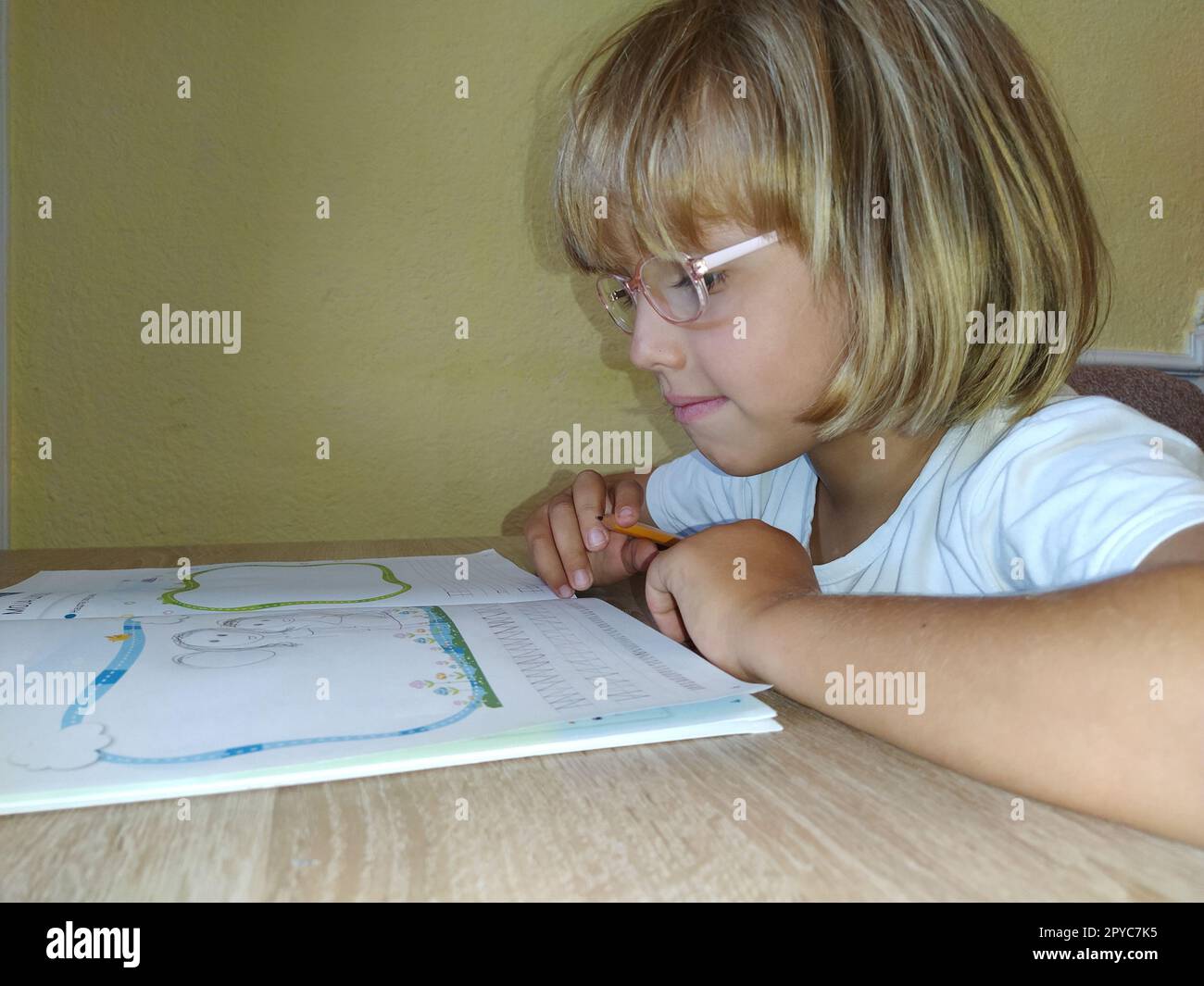 A 6-7 year old girl is reading a book or textbook and smiling. The child has blond hair, dark skin and glasses on his face. The child sits at the desk. yellow wall in the background Stock Photo