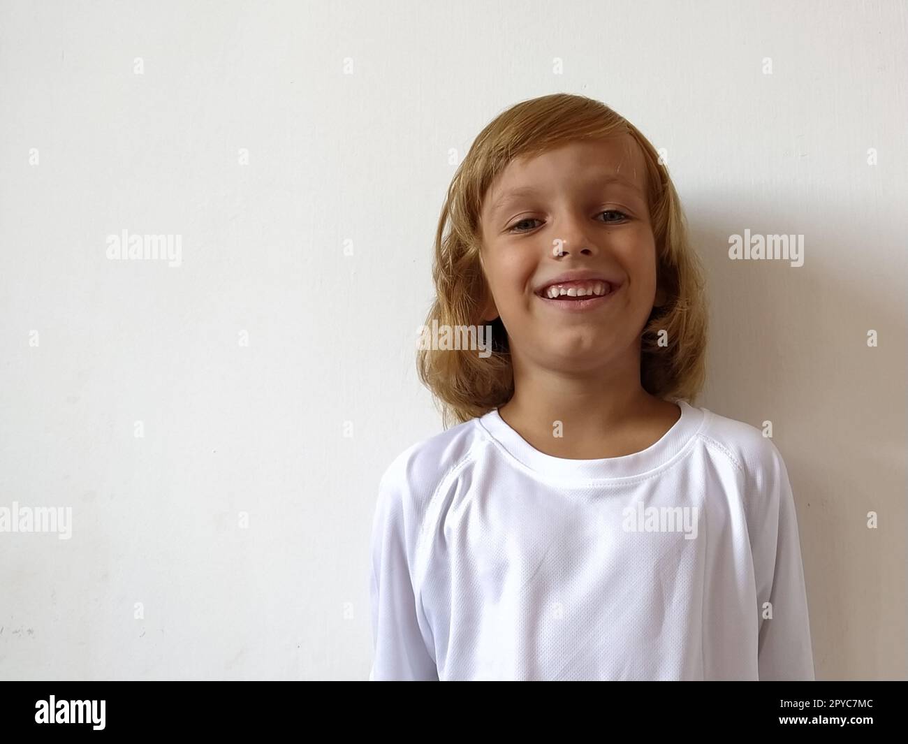 Little pretty girl age 6-7 years with light hair expresses emotional joy and a smile on a white background. A child with tanned skin. White T-shirt. Copy space Stock Photo