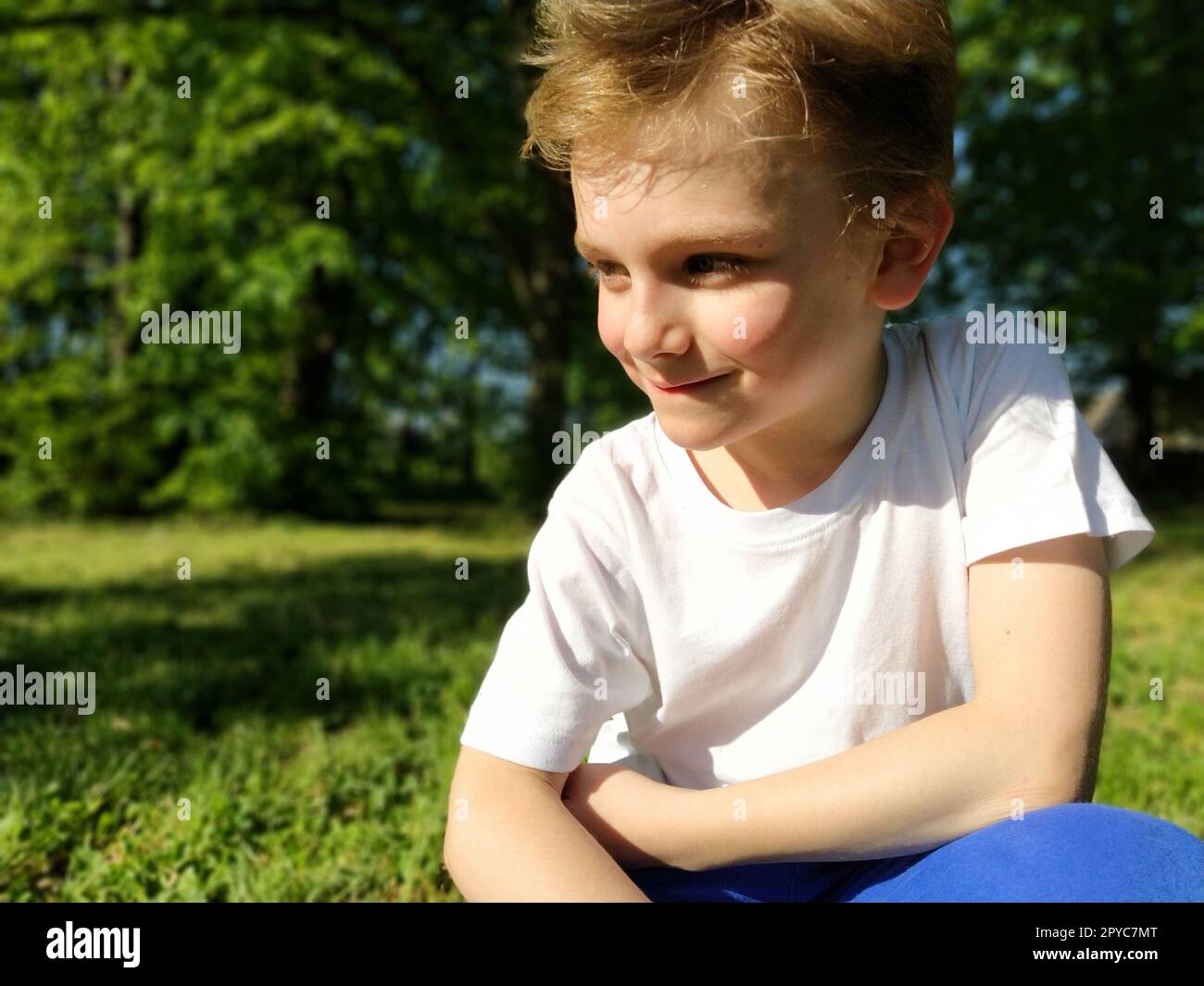 A cute young boy smiling and looks to the side. The child is dressed in a white T-shirt and blue pants. Blond hair is beautiful fall on the forehead. Long blonde eyelashes. Park in the background. Stock Photo