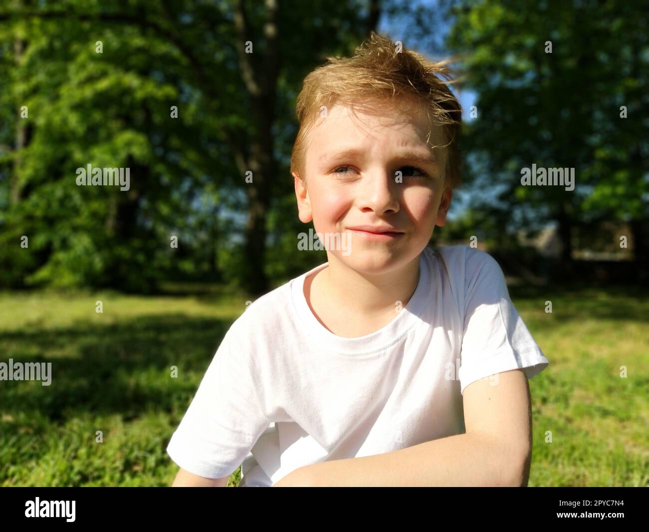 A cute young boy smiling and looks to the side. The child is dressed in a white T-shirt and blue pants. Blond hair is beautiful fall on the forehead. Long blonde eyelashes. Park in the background Stock Photo