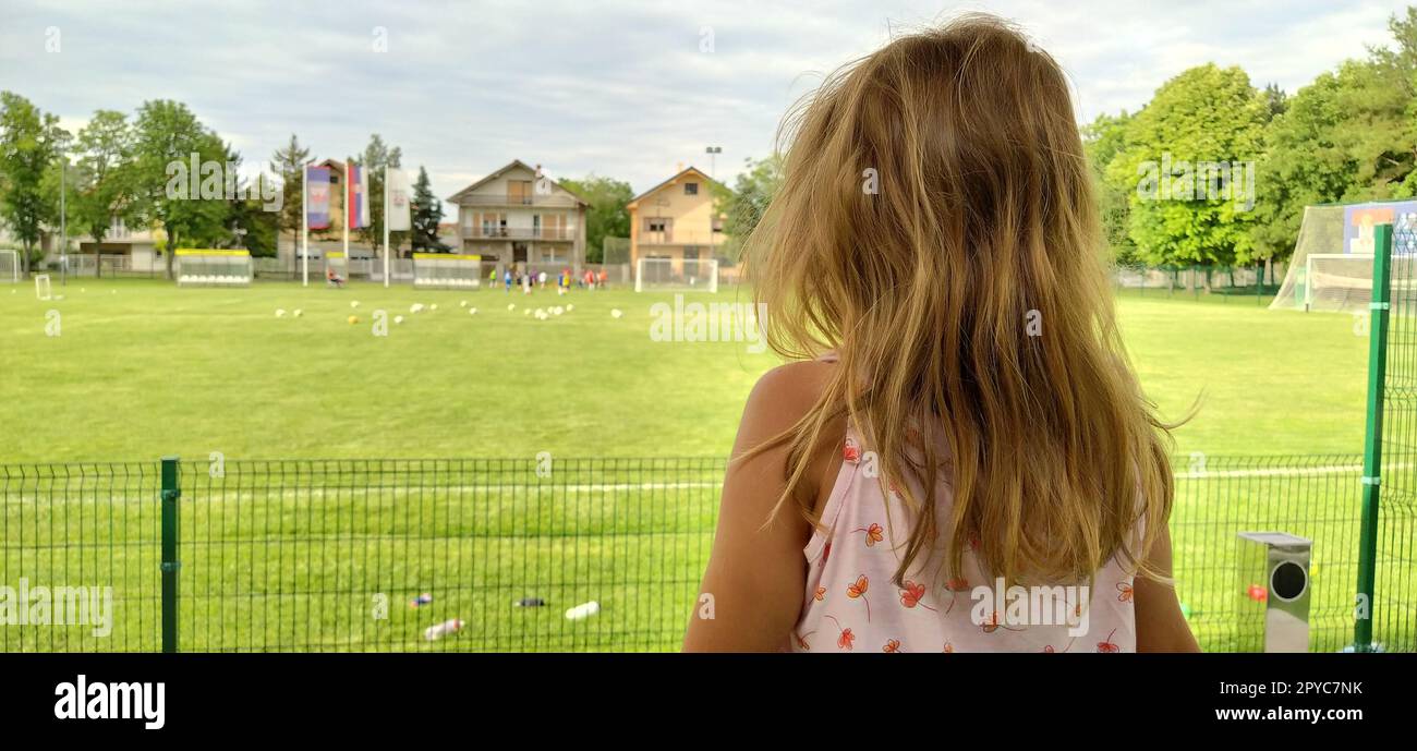 Sremska Mitrovica, Serbia, August 1, 2020. A girl sits with her back to the camera and watches a football match of childrens teams. Beautiful long blonde hair. Soccer fans in a match. Stock Photo