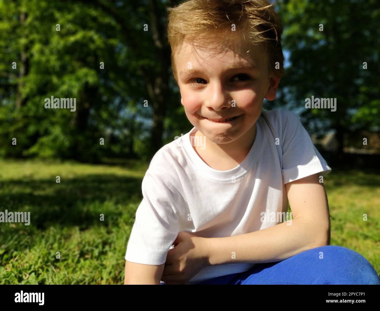 A cute young boy smiling and looks to the side. The child is dressed in a white T-shirt and blue pants. Blond hair is beautiful fall on the forehead. Long blonde eyelashes. Park in the background. Stock Photo