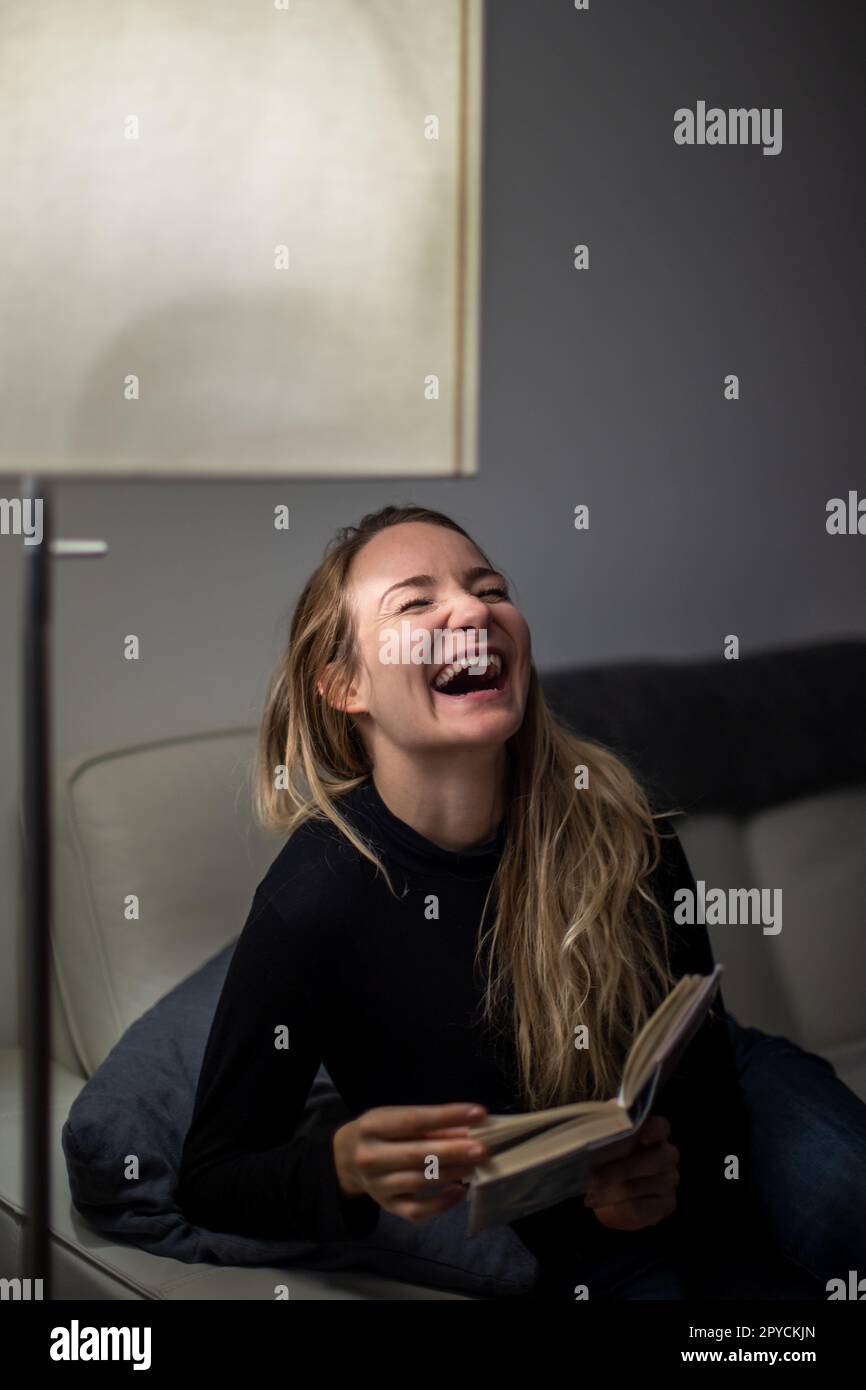 Avid reader laughing. Young attractive girl happy positive smile sit sofa home relax read book story novelty laugh humor joke Stock Photo