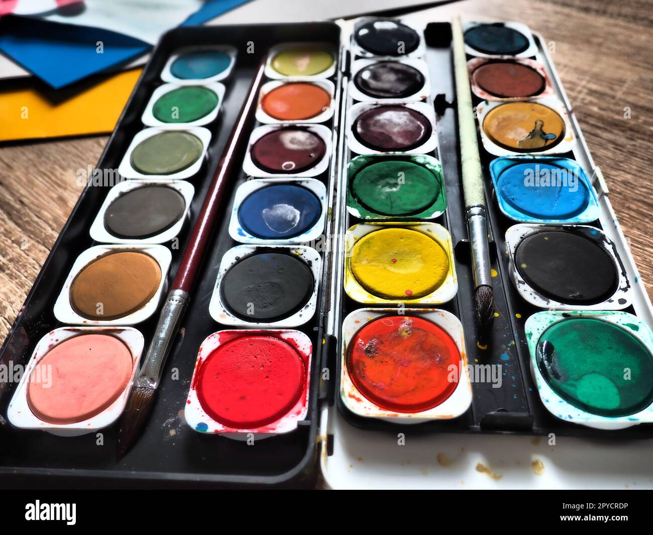 A set of watercolors of different colors for drawing. Multicolored round containers with bright paint. Tools for the lesson of drawing and labor. Colored paper. Red, blue, yellow, green colors. Stock Photo