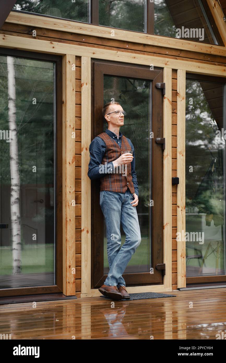 Pensive middle age man stands on the terrace of a wooden bungalow and drinks whisky Stock Photo