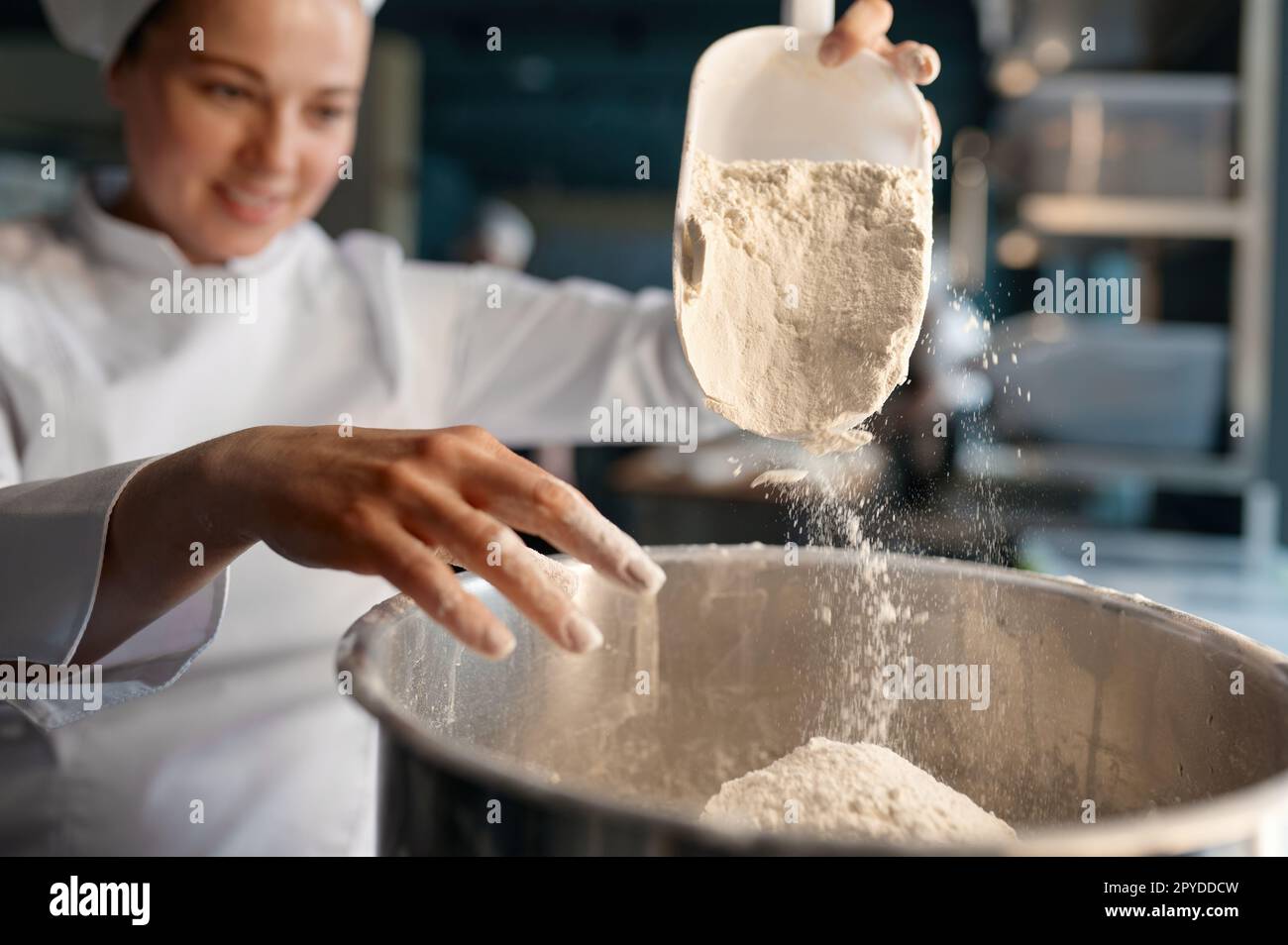 Female confectioner wearing white uniform putting flour into big metal bowl Stock Photo