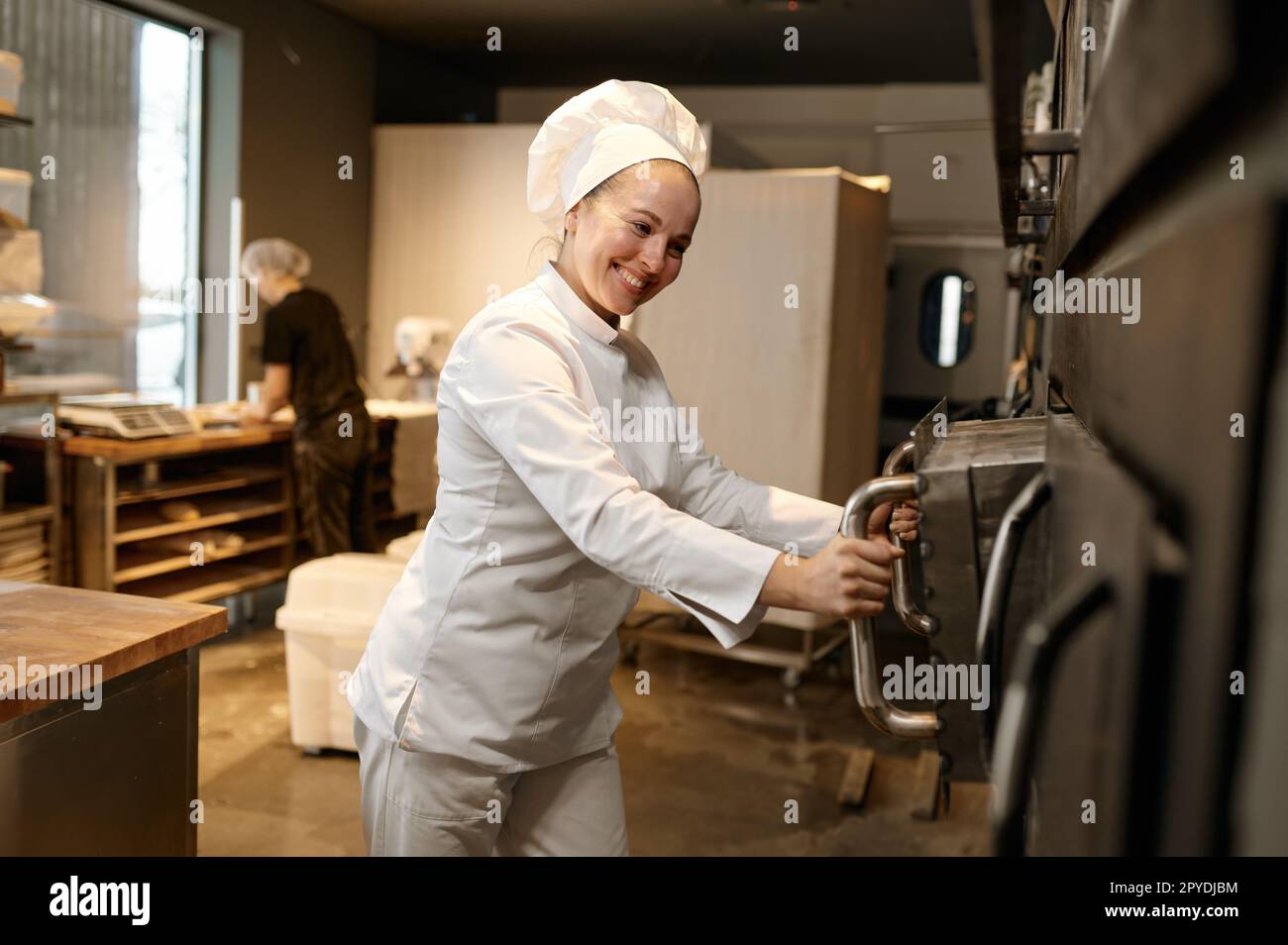 Smiling woman baker opening big industrial oven at modern bakery ...