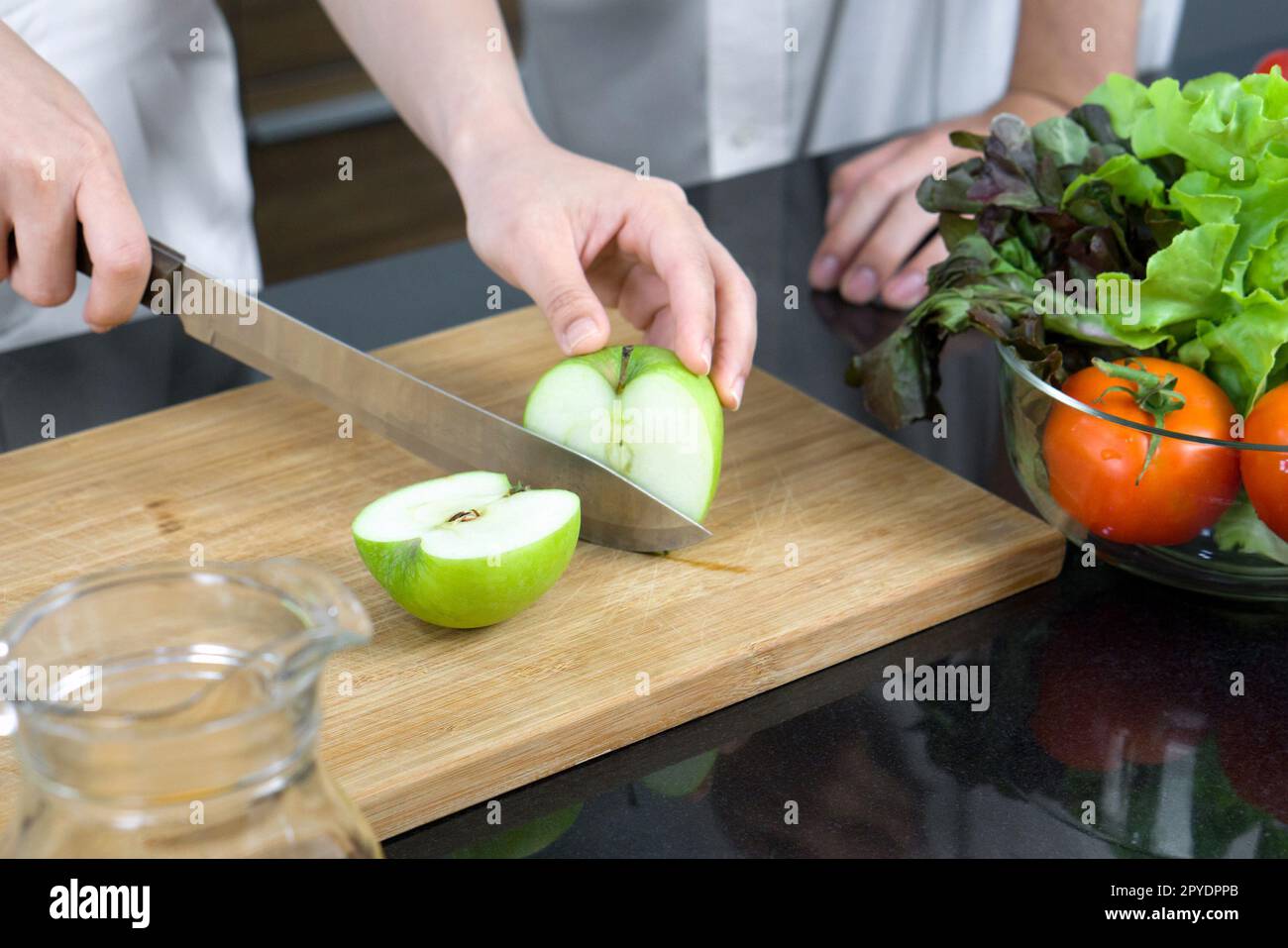 Closeup hand holding knife cutting green apple on a wooden chop board. A glass bowl with a variety of vegetables is placed on the kitchen counter. Stock Photo