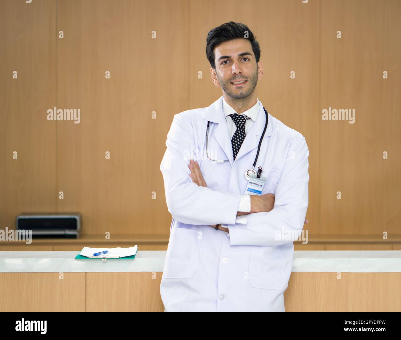 Portrait of handsome doctor with stethoscope wearing white gown uniform standing in front of ward counter in the hospital. Stock Photo