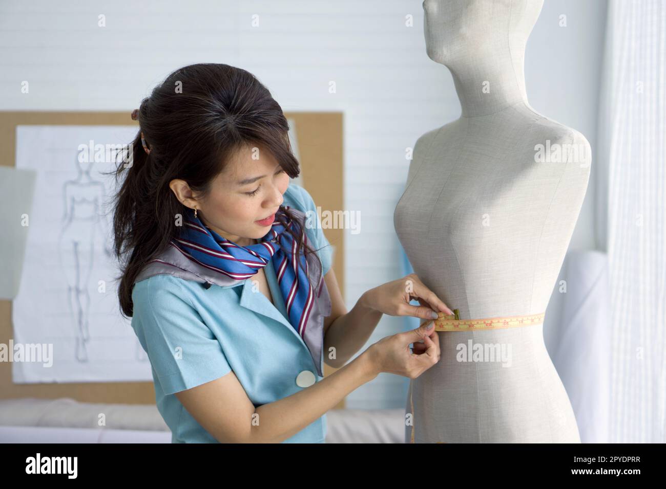 Fashion designer room. Young asian dressmaker in striped scarf use tape measure, measuring waist size of the mannequin. Stock Photo