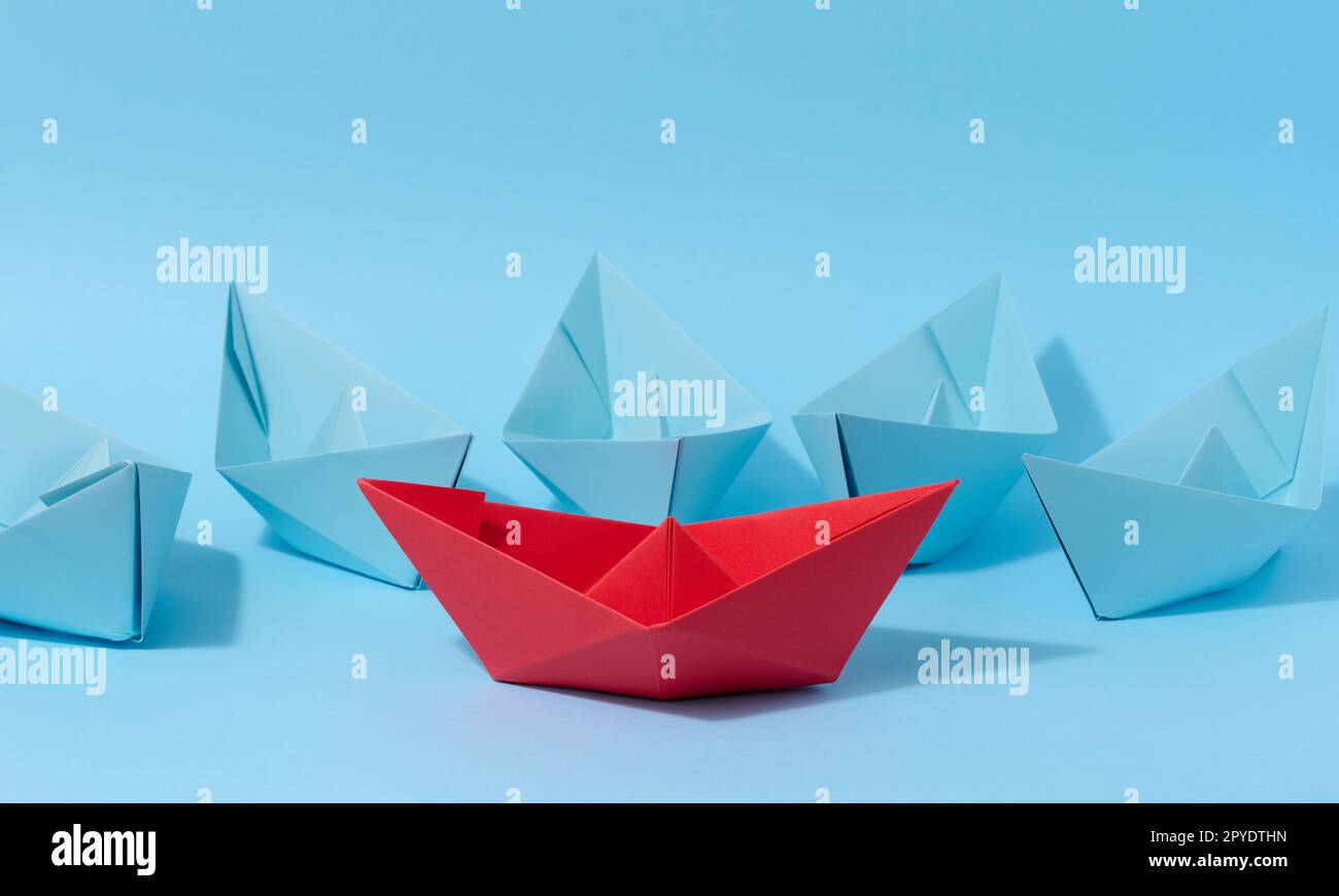 A red paper boat stands in front of a group of blue paper boats, a confrontation Stock Photo