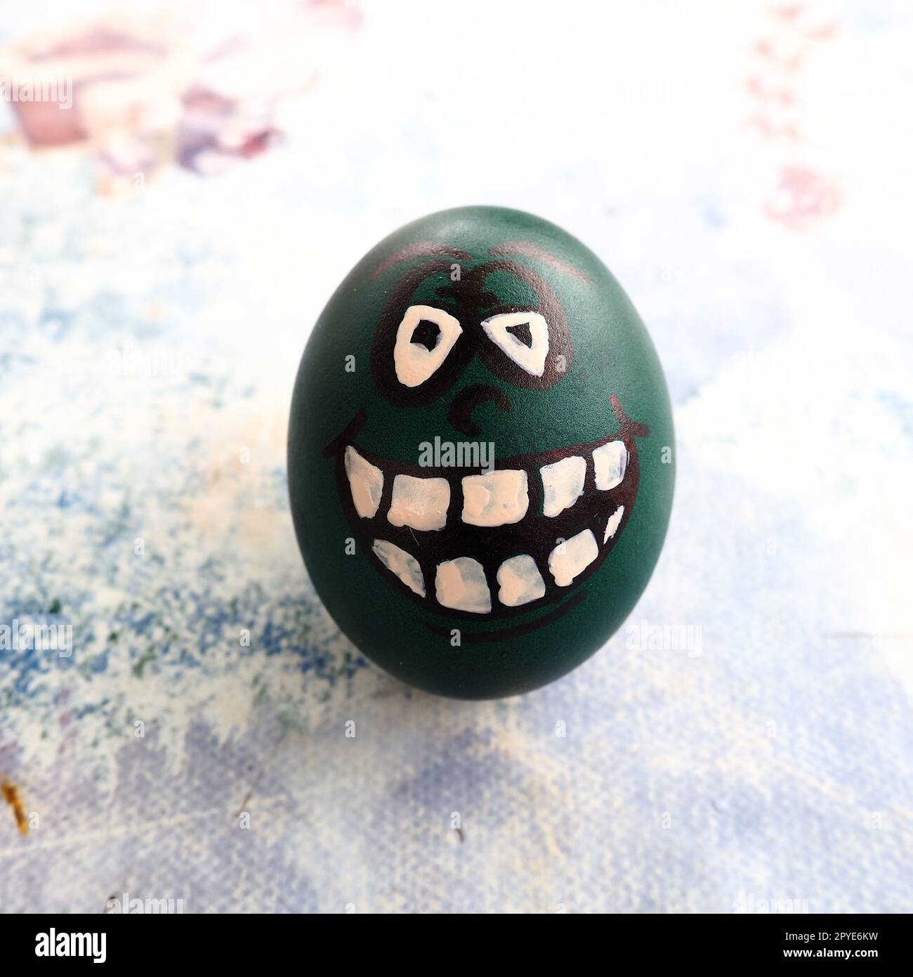 Easter egg dyed green with a painted laughing face. Cool grimace with eyes and big white teeth. Scary mug for Halloween. Emoticon for Easter. Light abstract background. Stock Photo