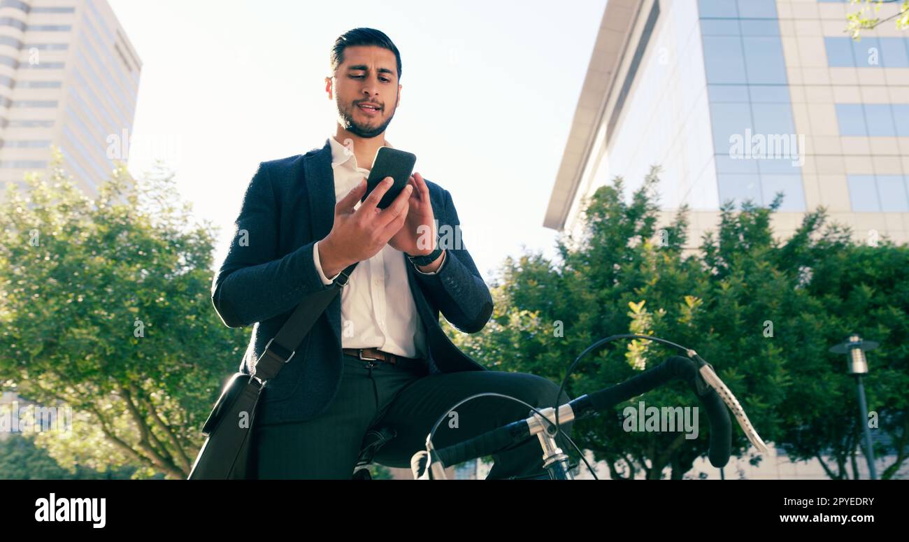 Sending a few message on his way to work. Low angle shot of a handsome young businessman using his cellphone while commuting to work on his bicycle. Stock Photo