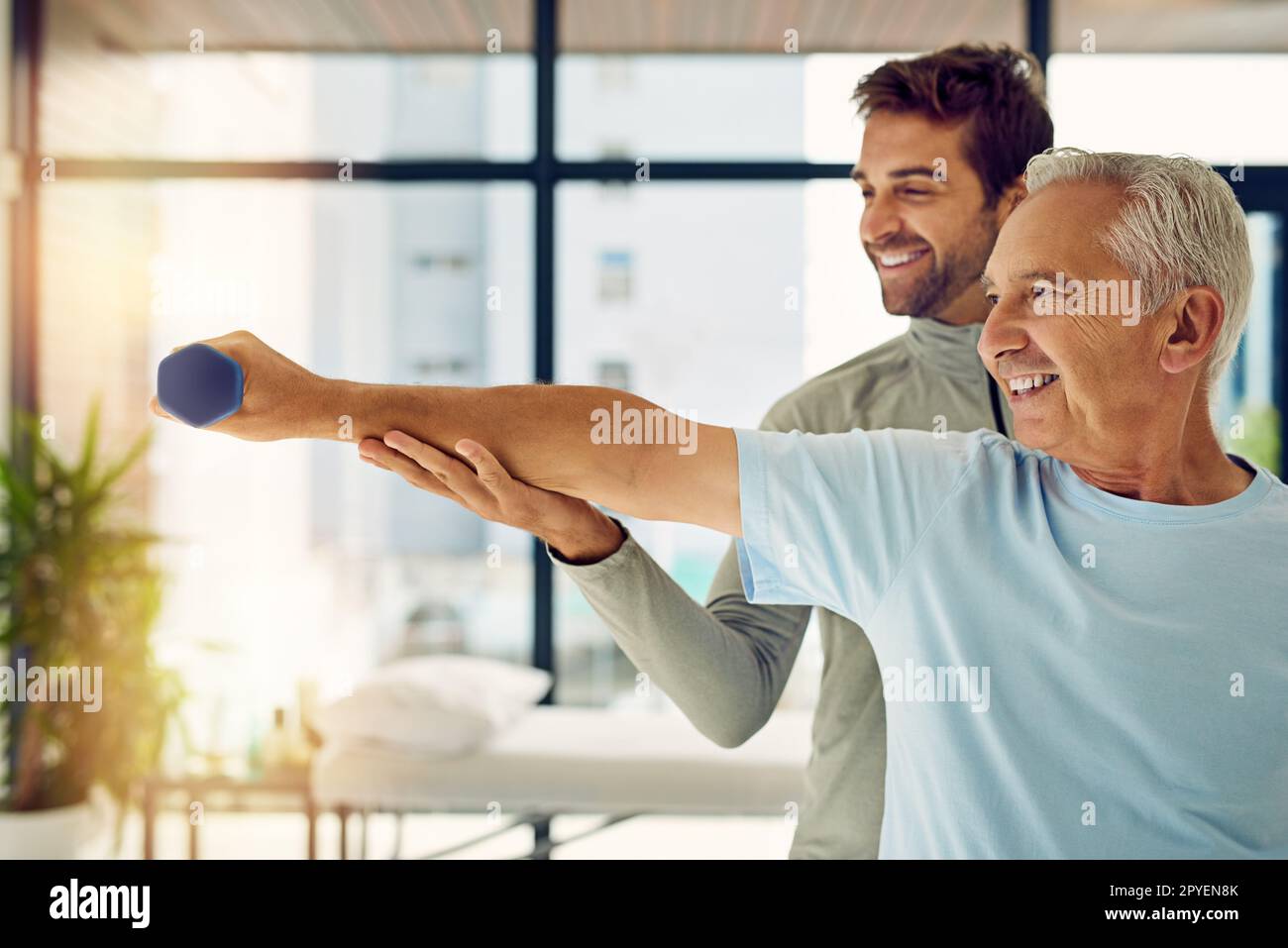He has a companion on the road to fitness. a friendly physiotherapist helping his senior patient work out with weights. Stock Photo
