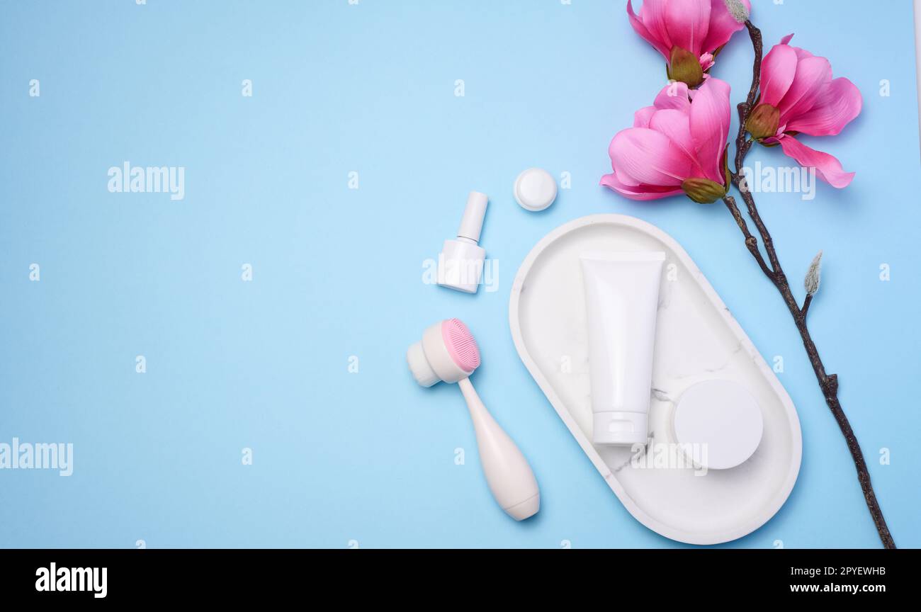 White plastic tubes and jars of cream, and a massage brush for facial cleansing on a blue background, items for cosmetic procedures Stock Photo