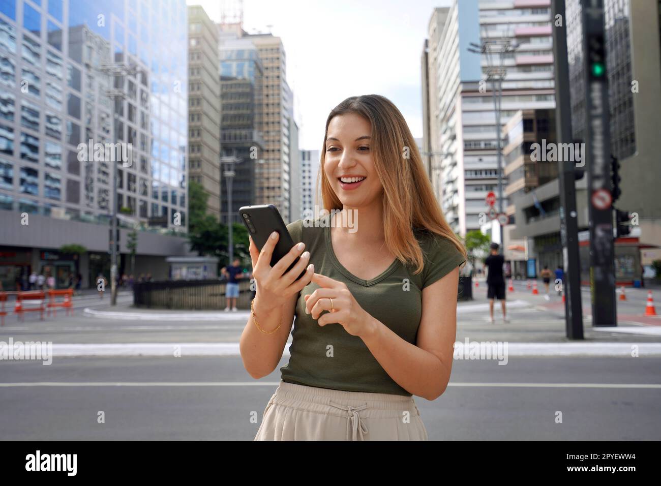 Cheerful Brazilian girl watching funny video on phone standing on street at daytime. Brown-haired with toothy smile wears green t-shirt. Social media addiction concept. Stock Photo