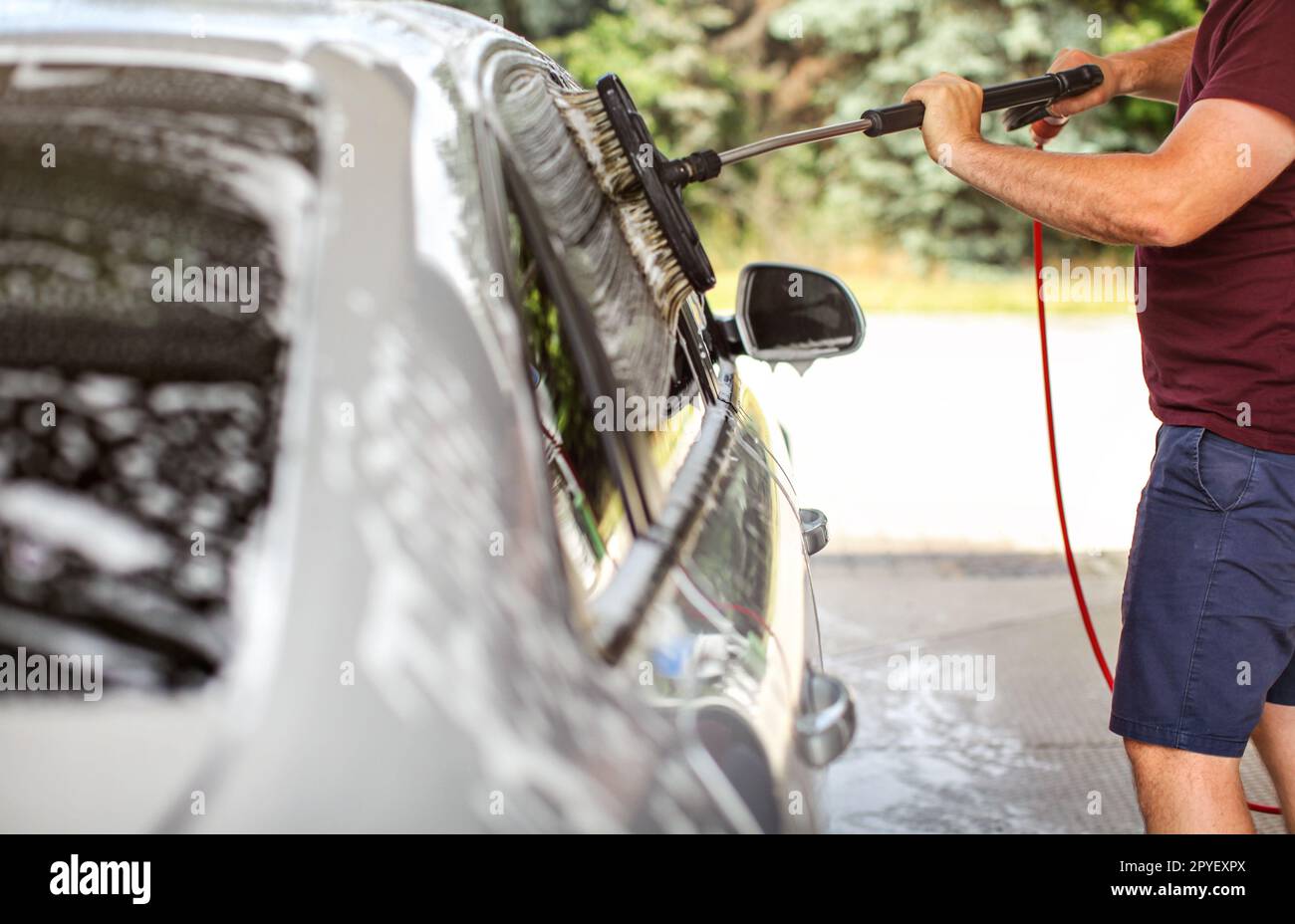 Young man in shorts and t shirt washing his car in self serve carwash, cleaning side windows with brush. Stock Photo