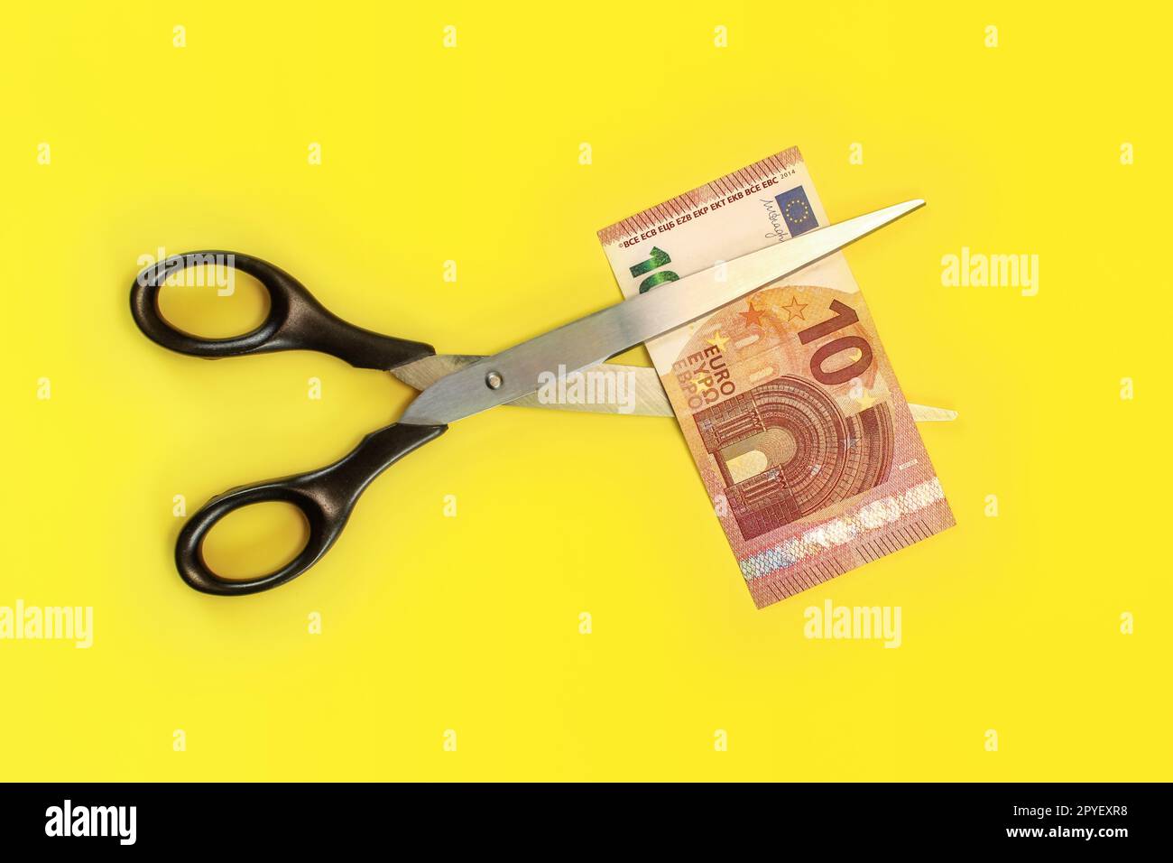 Tabletop view - large scissors cutting ten eur banknote over yellow board. Cut fees concept. Stock Photo